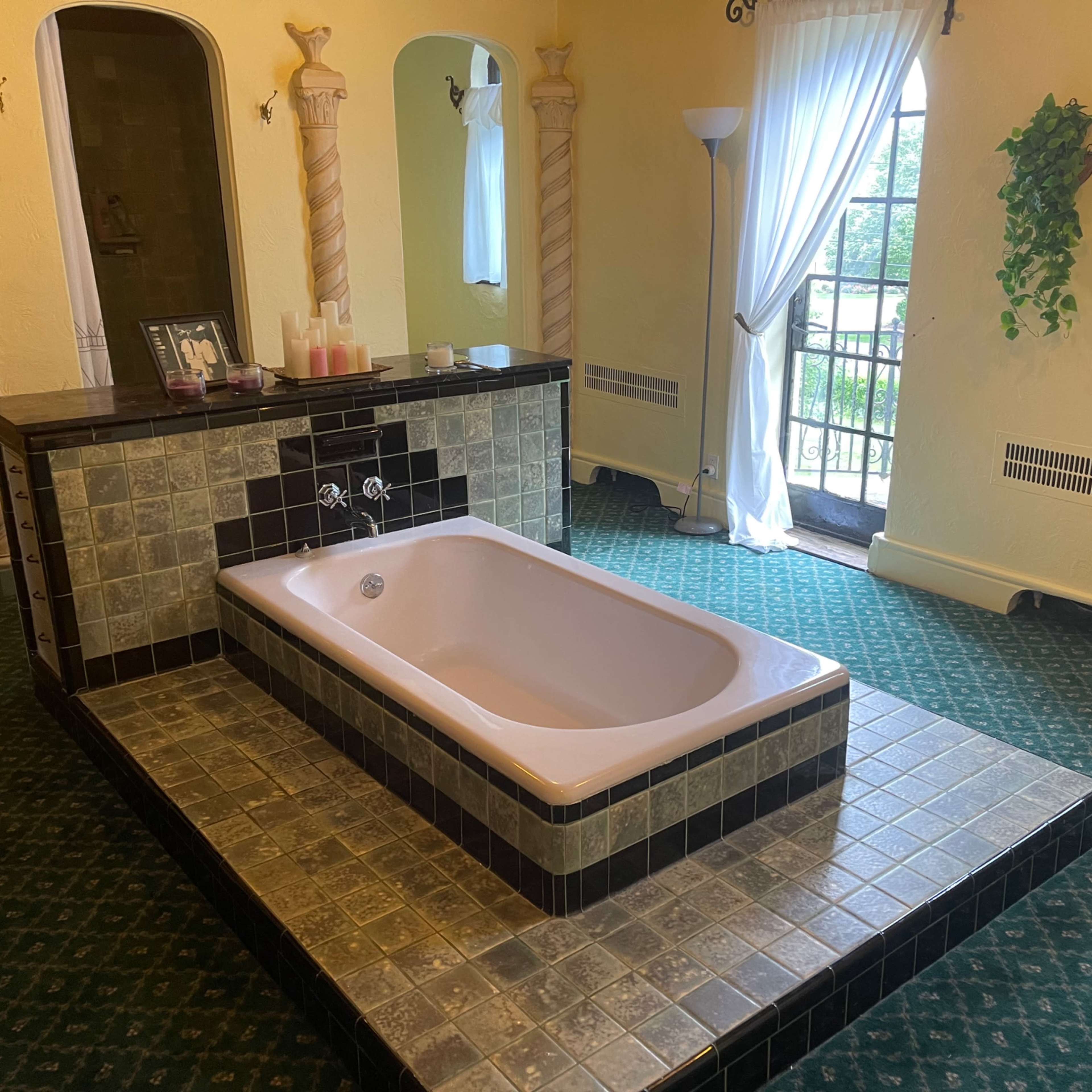 A bathroom featuring a large pink bathtub set on a tiled platform, with decorative columns and a window overlooking a balcony.