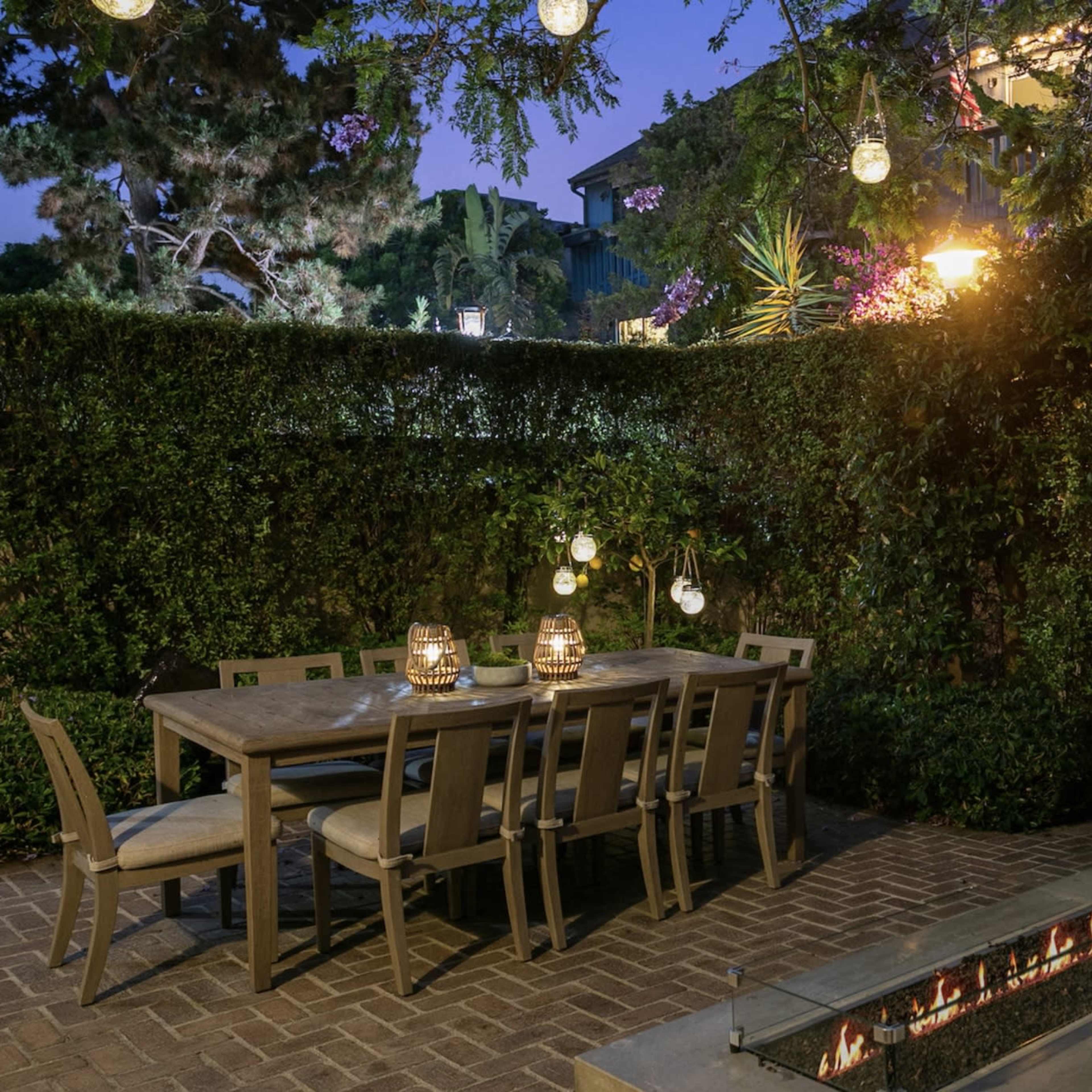 A dining area with a large table and chairs, surrounded by greenery and illuminated by hanging lights, features a firepit nearby.