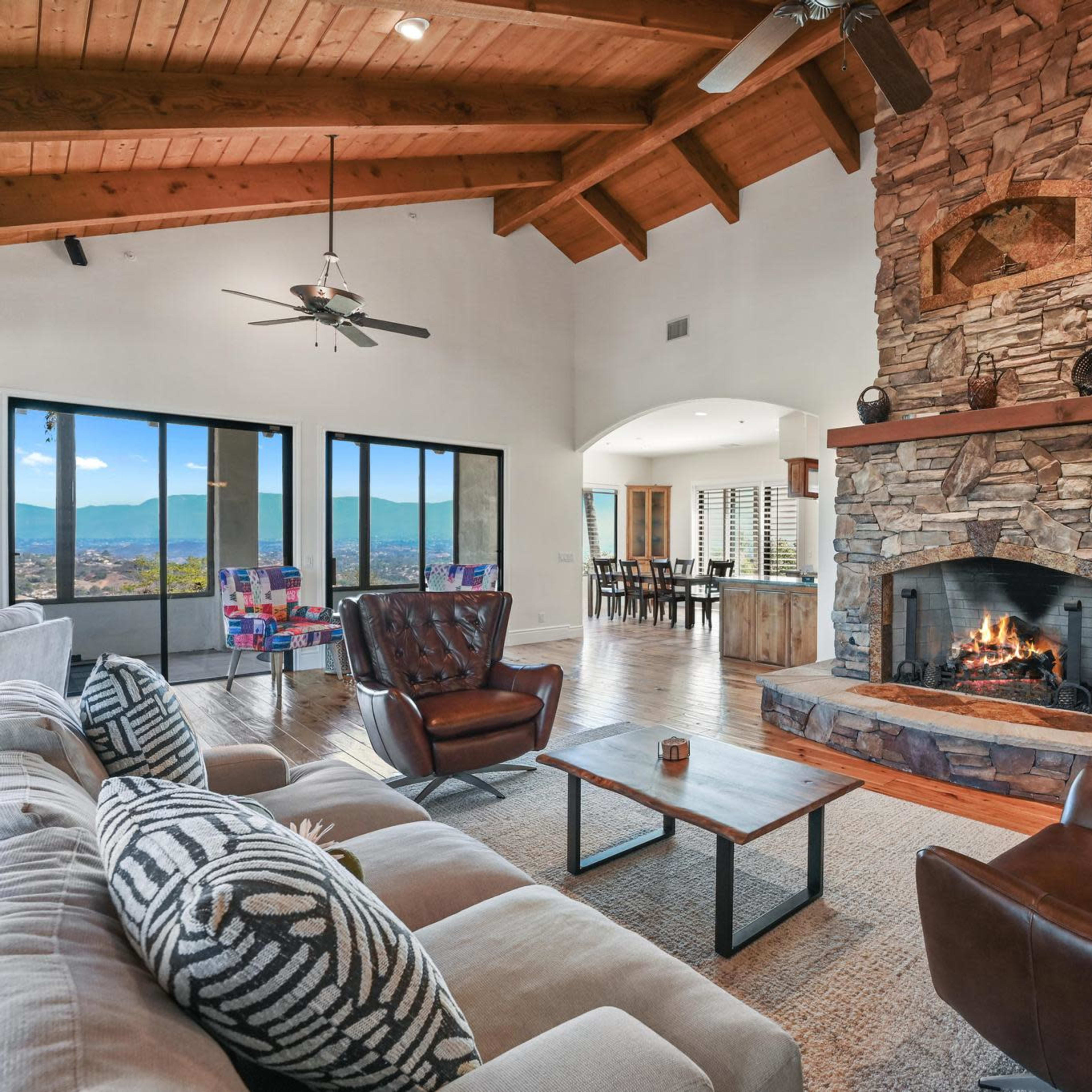 A spacious living room features a stone fireplace, wooden beams, large windows with mountain views, and comfortable seating arrangements.