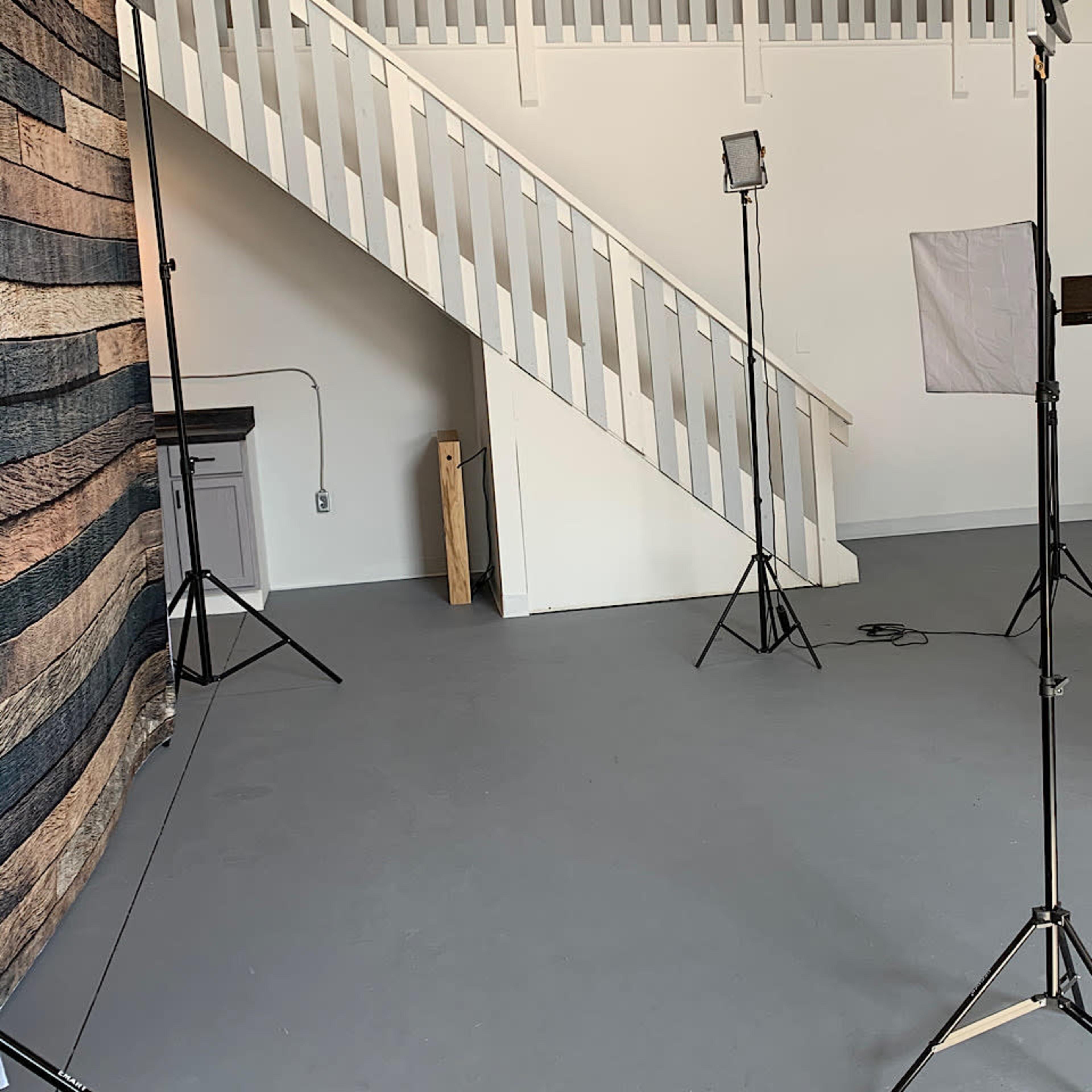 A photography studio setup with several light stands, soft boxes, and a wooden backdrop, alongside a staircase and a white wall.
