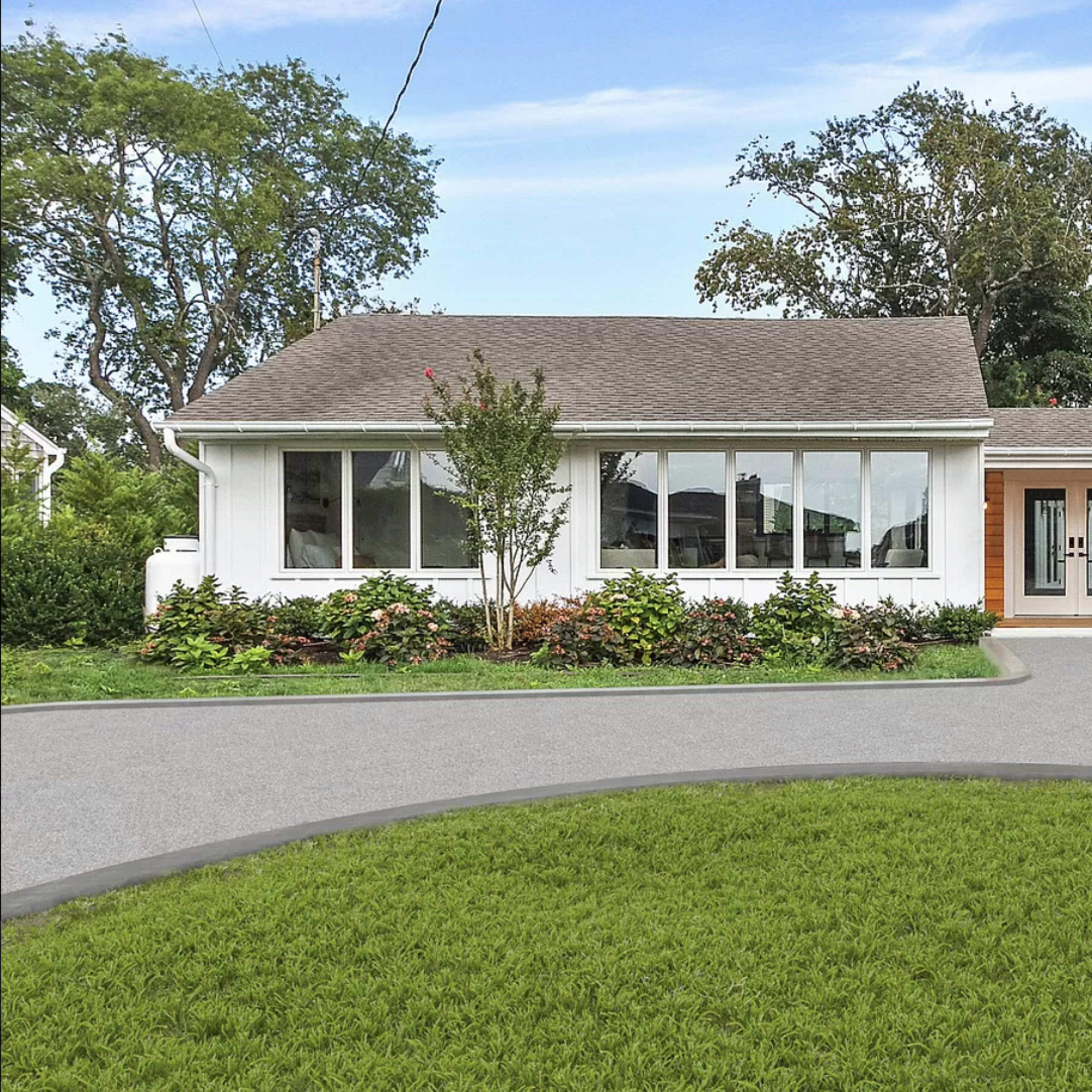 A single-story house with large front windows and a wooden door is surrounded by landscaped greenery and a curved driveway.