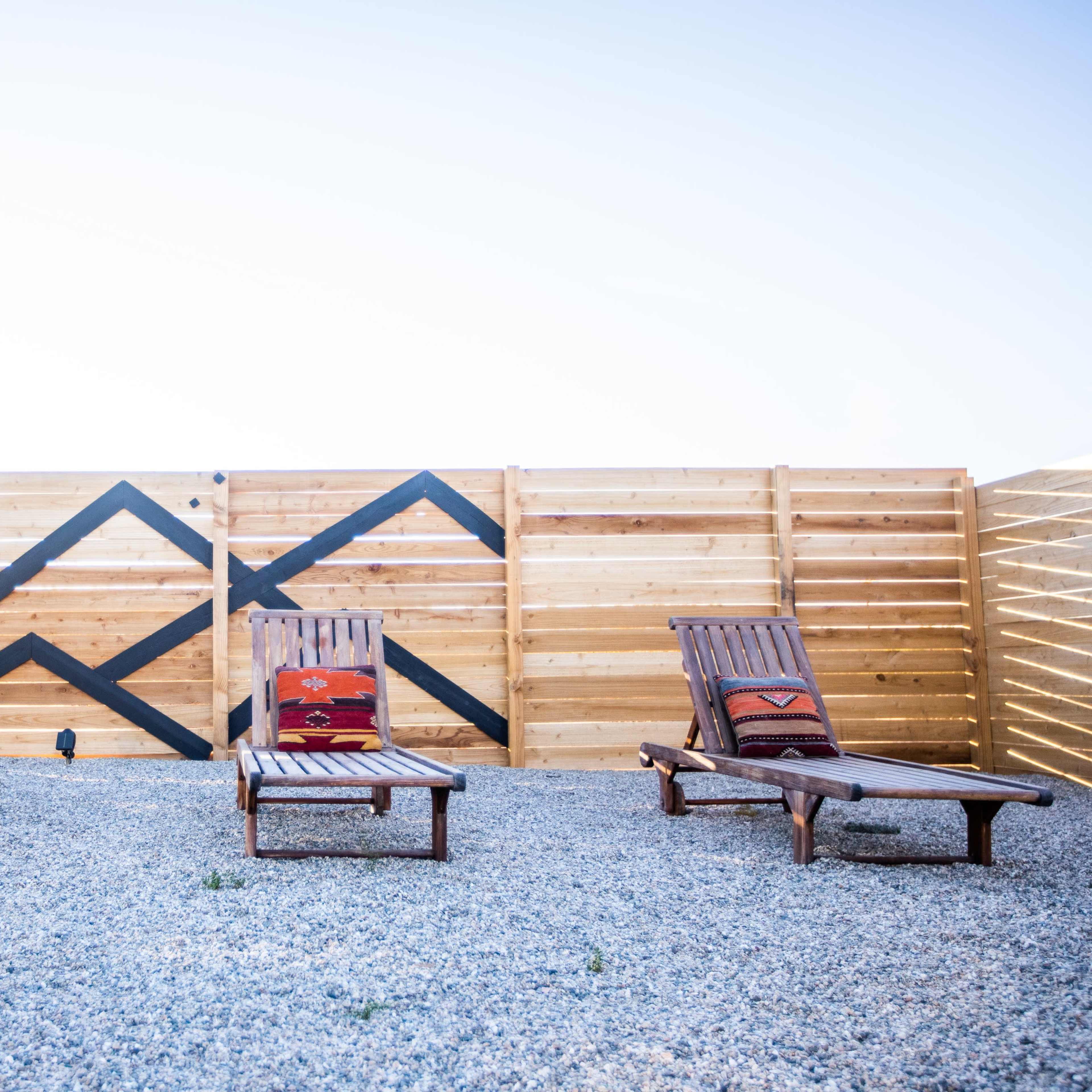 Three wooden lounge chairs with patterned cushions sit on a gravel patio in front of a wooden fence featuring a geometric design.