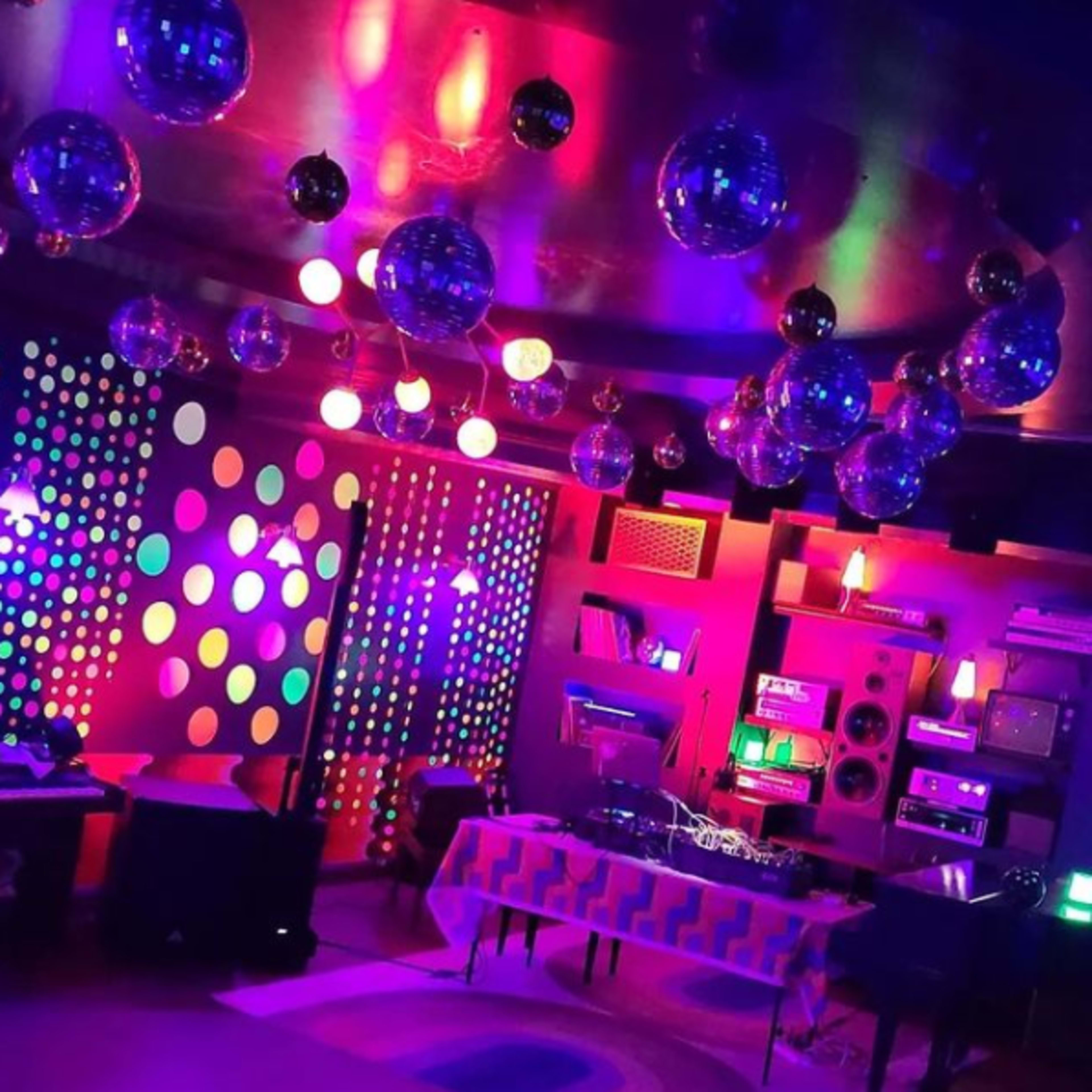 A vibrant retro-themed room adorned with colorful disco balls and polka-dotted walls, equipped with music equipment and ambient lighting.