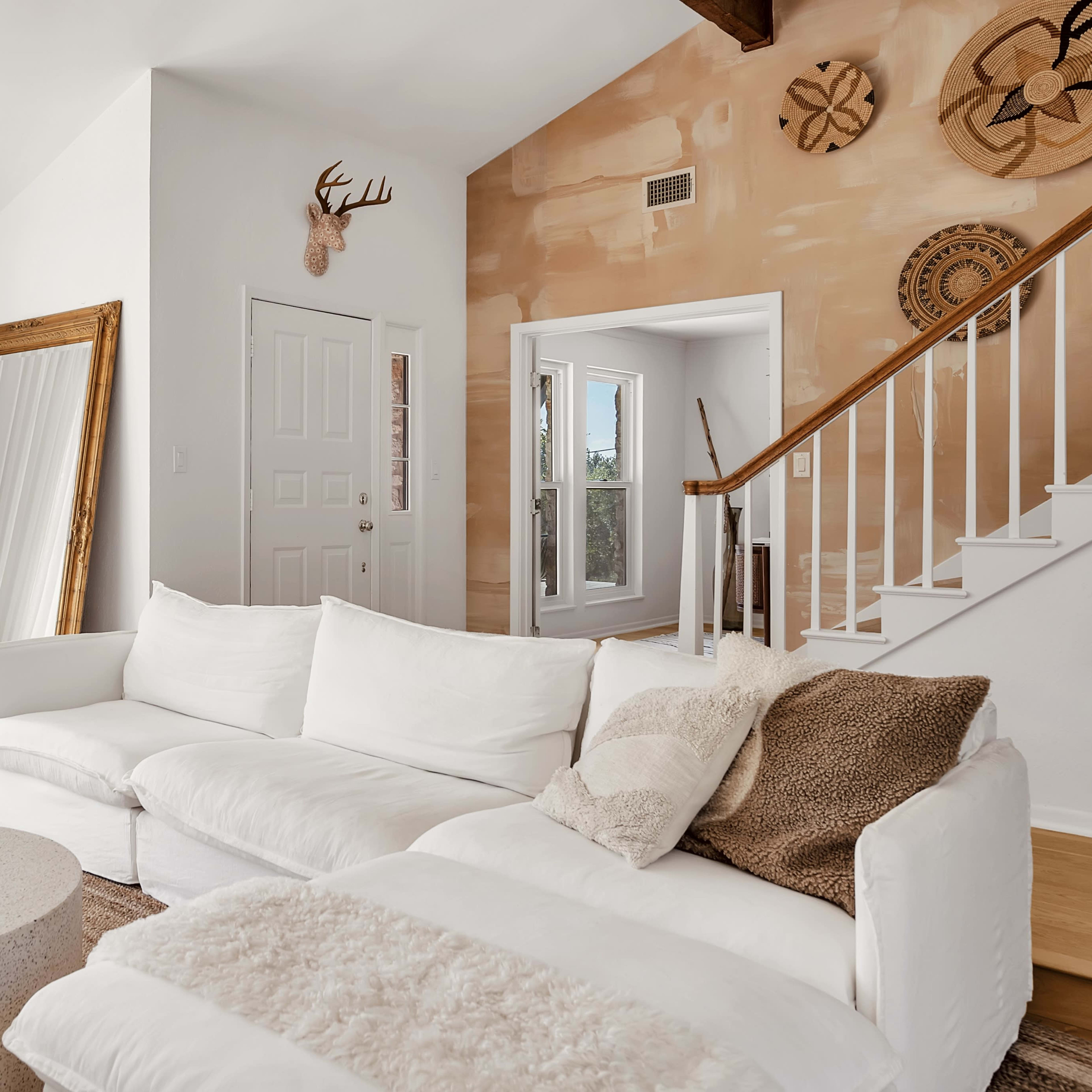 The living room features a white sectional sofa, a round coffee table, large windows, and a staircase leading to another area.