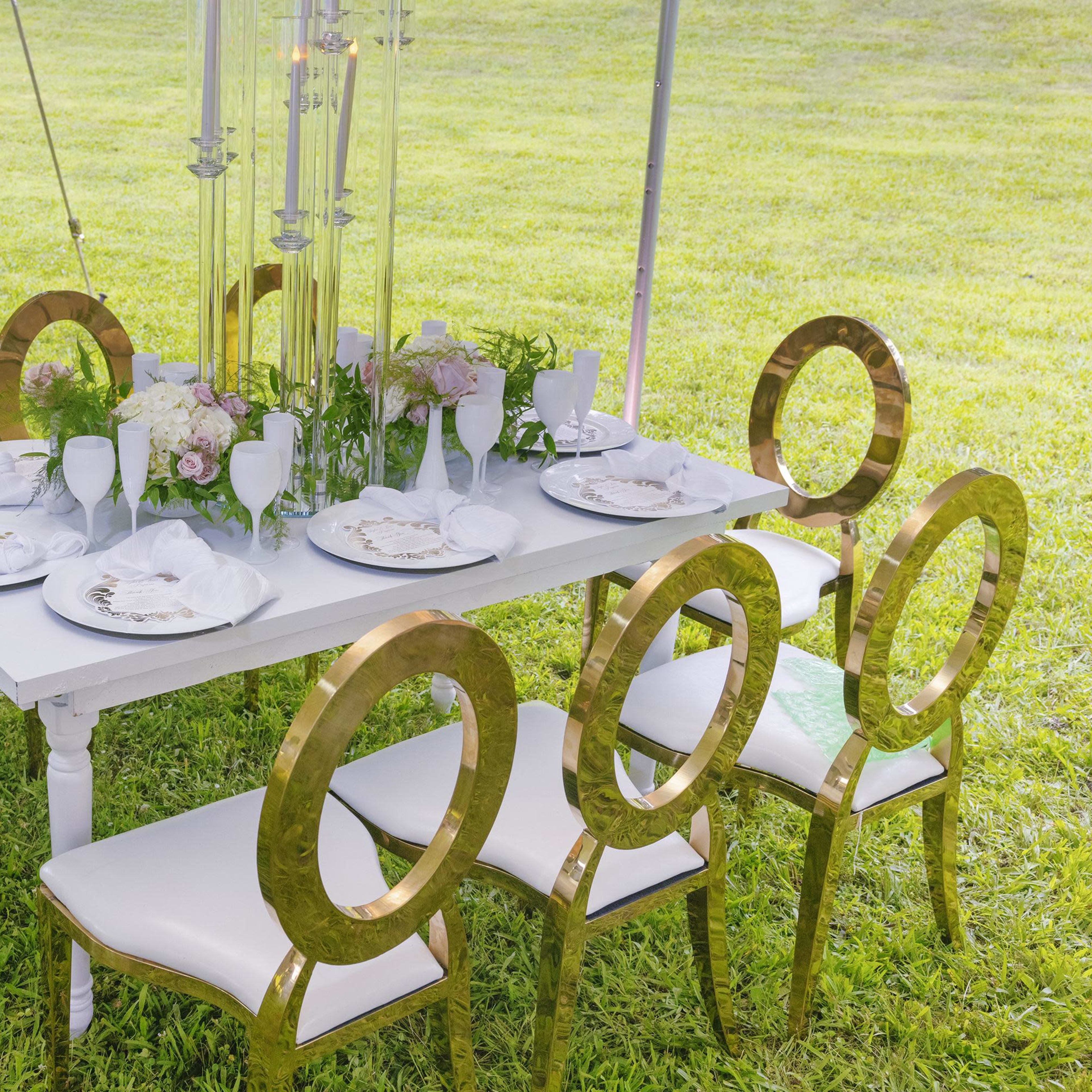A white table is set with plates and floral arrangements, surrounded by gold-accented chairs on a grassy area.