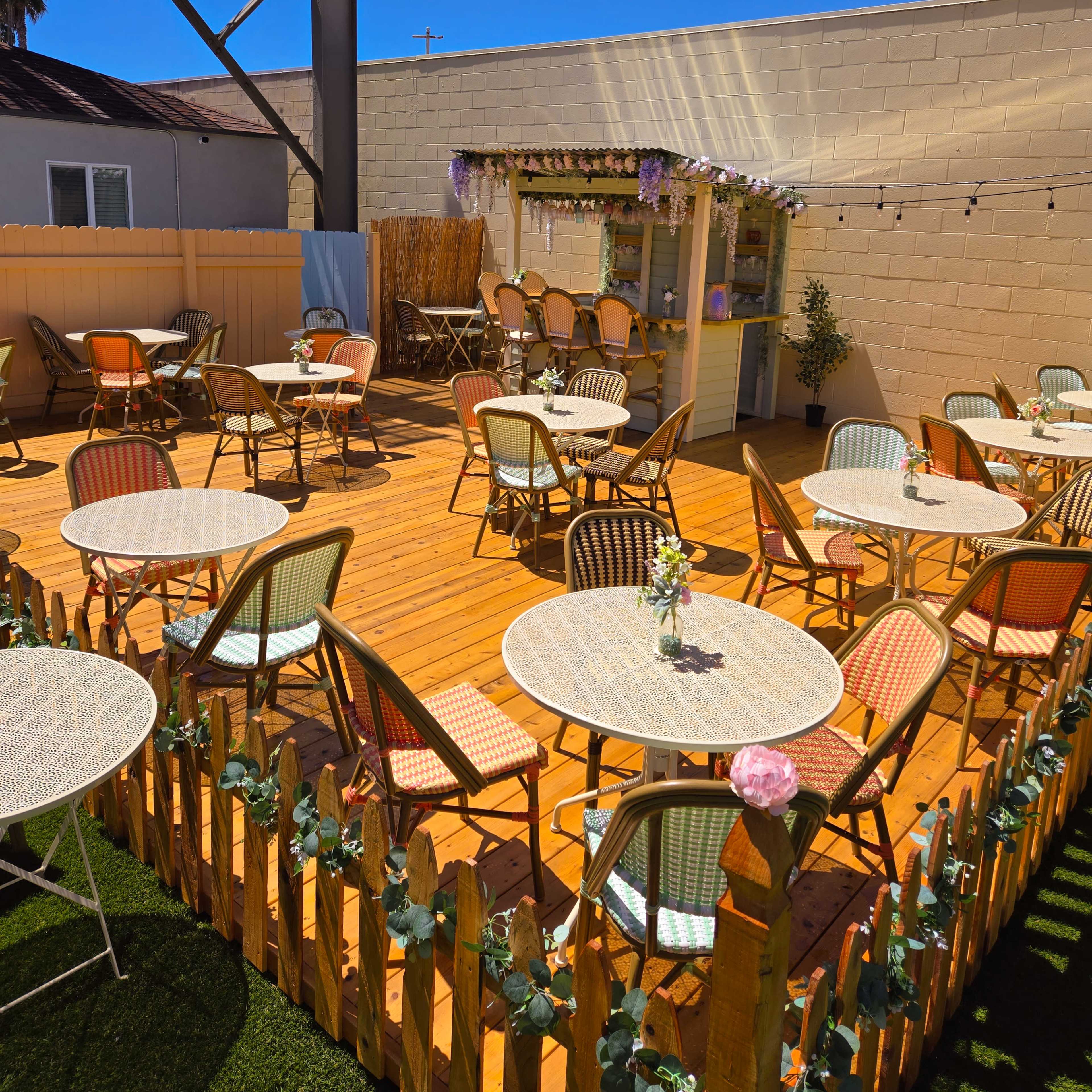 A wooden patio area with several round tables and chairs, surrounded by a low fence and decorated with string lights and flowers.