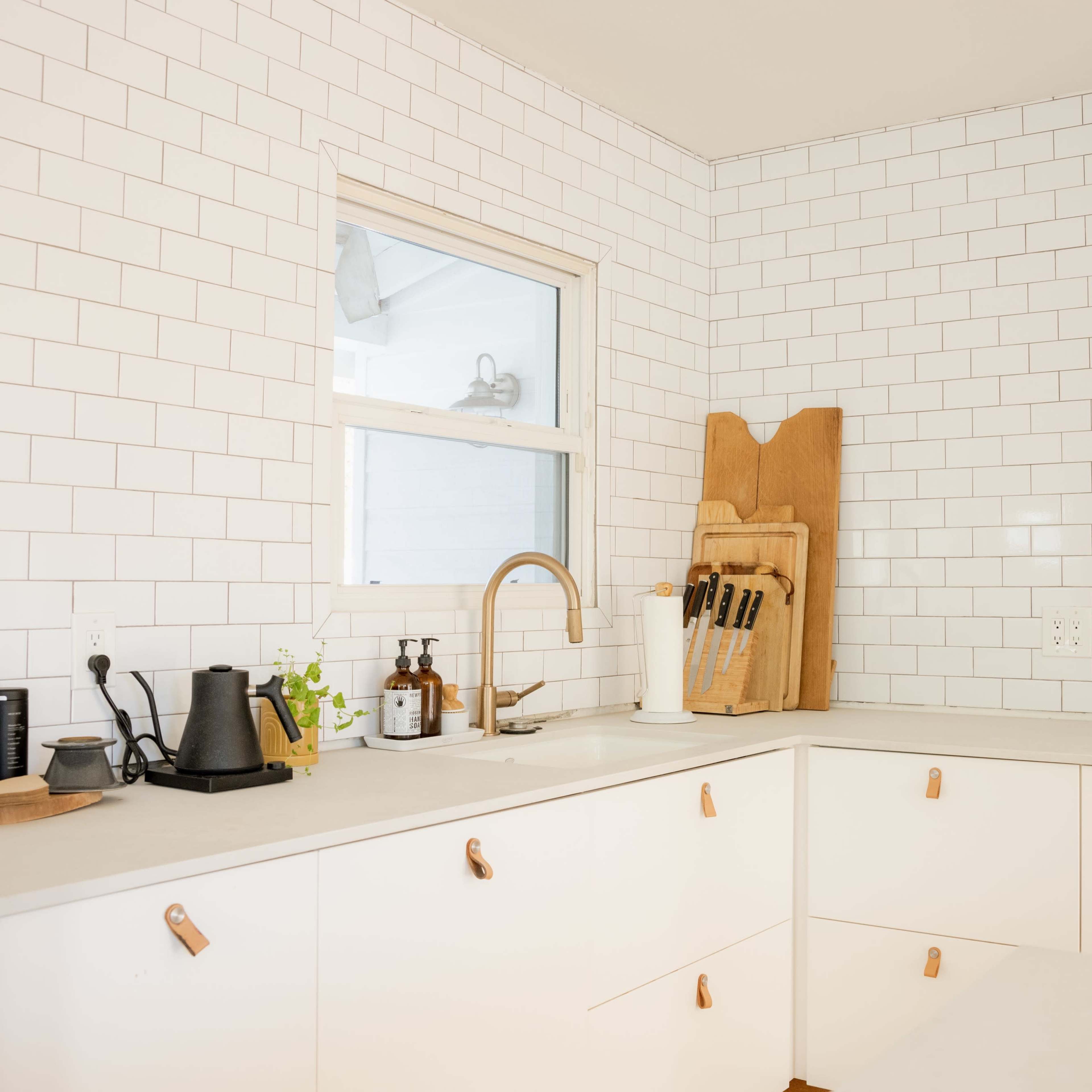 A modern kitchen features white subway tile walls, a corner sink with a faucet, and minimalistic cabinetry with wooden accents.