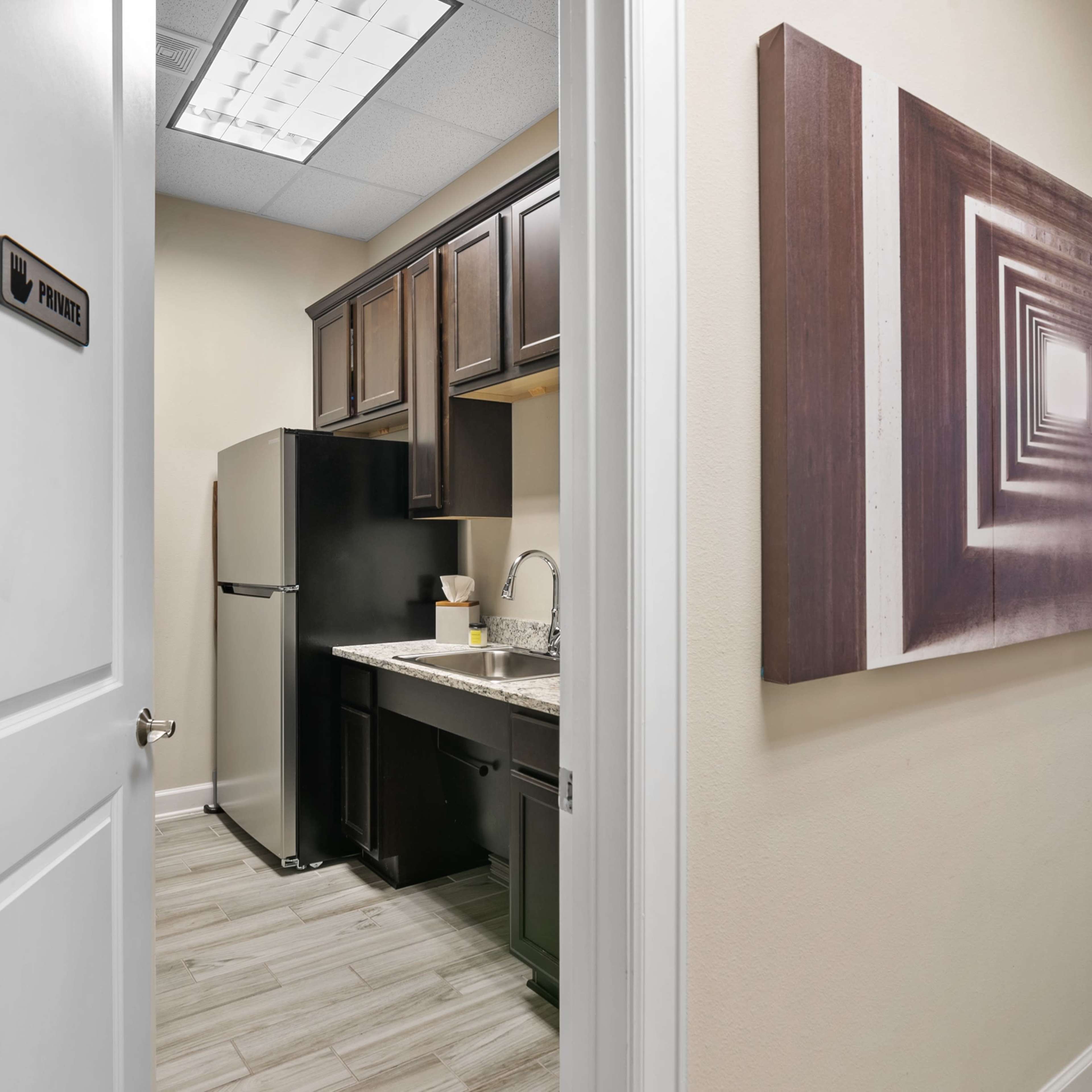 A door opens to reveal a compact kitchen area with dark cabinets and a refrigerator, accompanied by a geometric artwork on the wall.