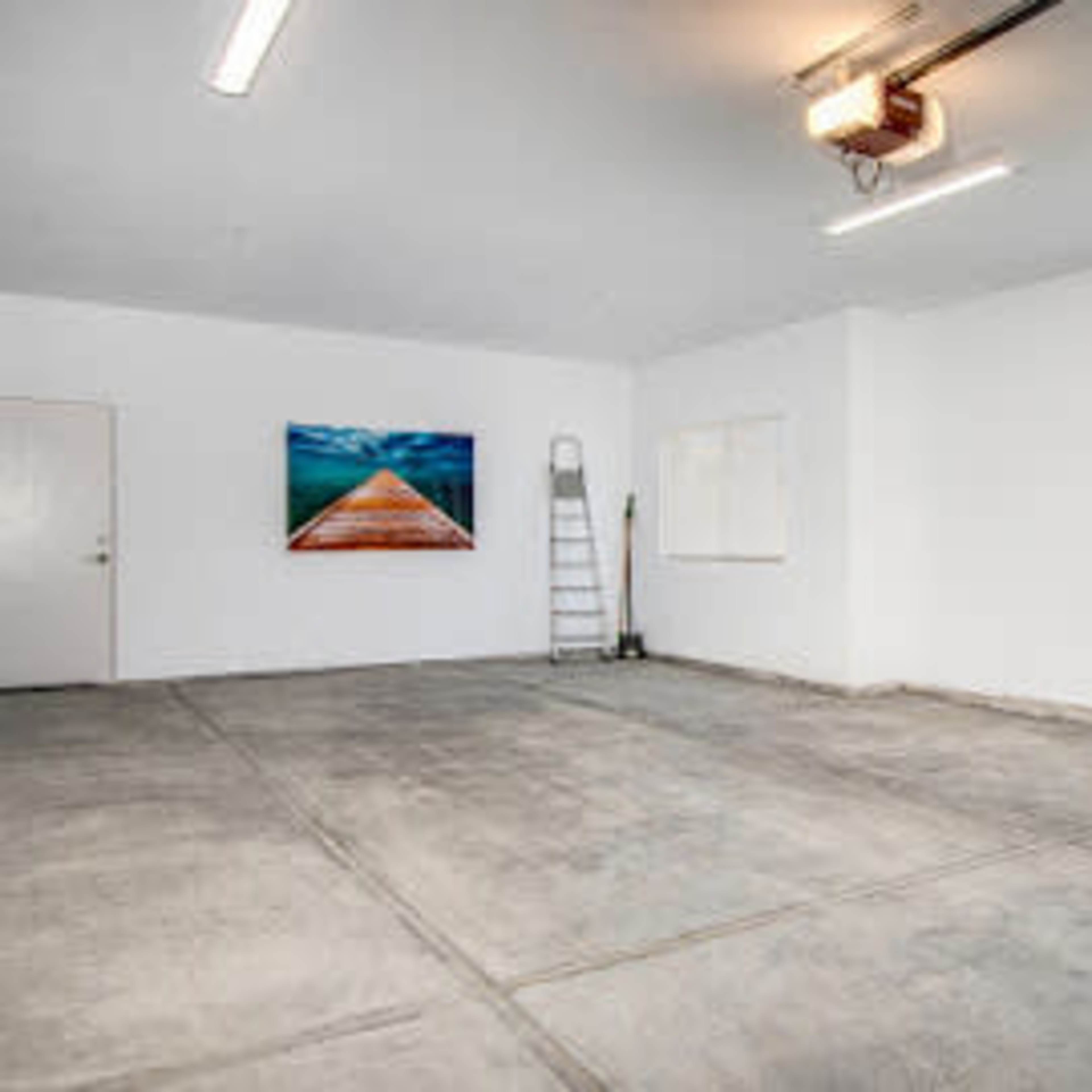 A clean, empty garage with a concrete floor, a door, two windows, a ladder, and a large photo of a pier on the wall.