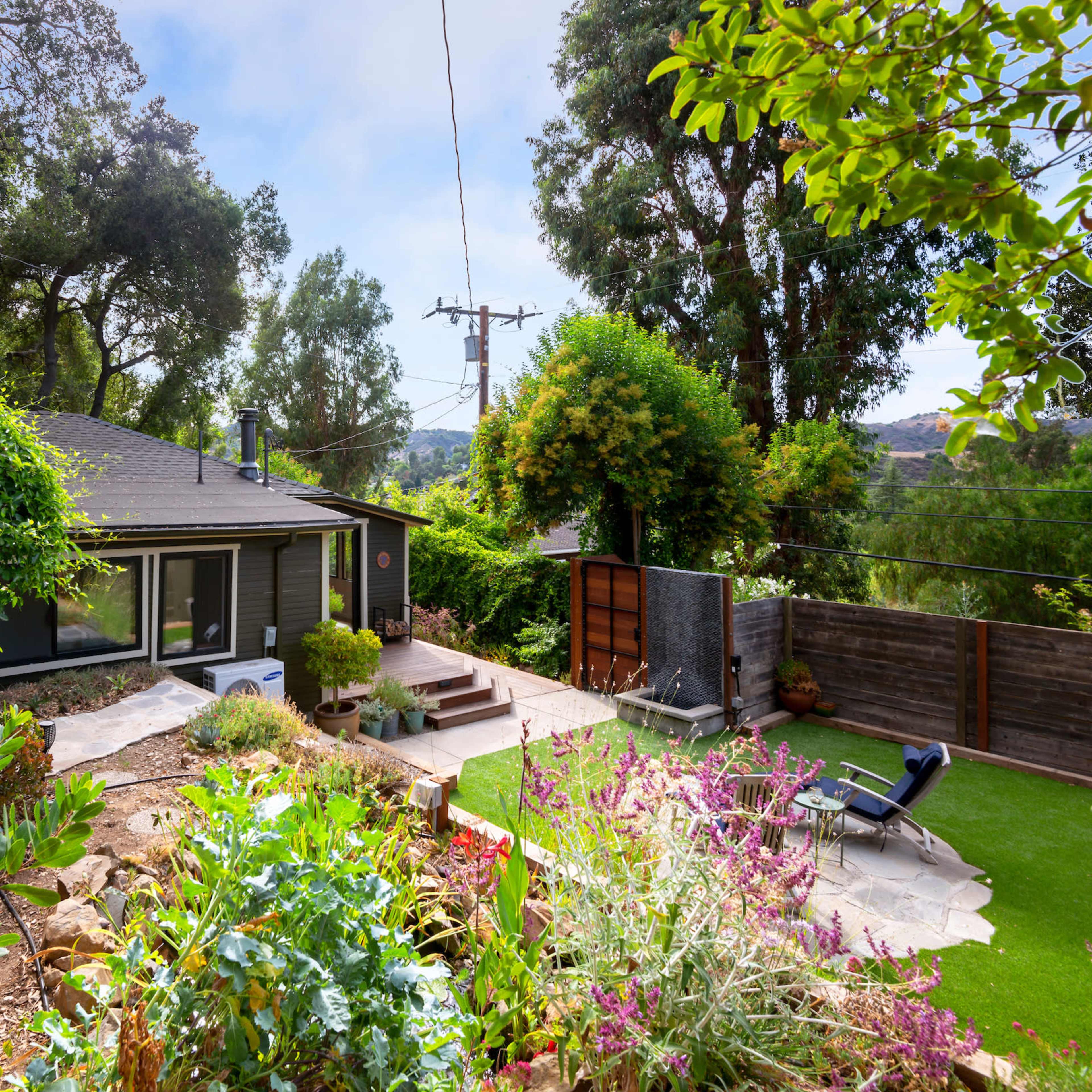 A small house is surrounded by lush greenery and colorful flowers, with a stone pathway leading to its entrance and a lawn area nearby.