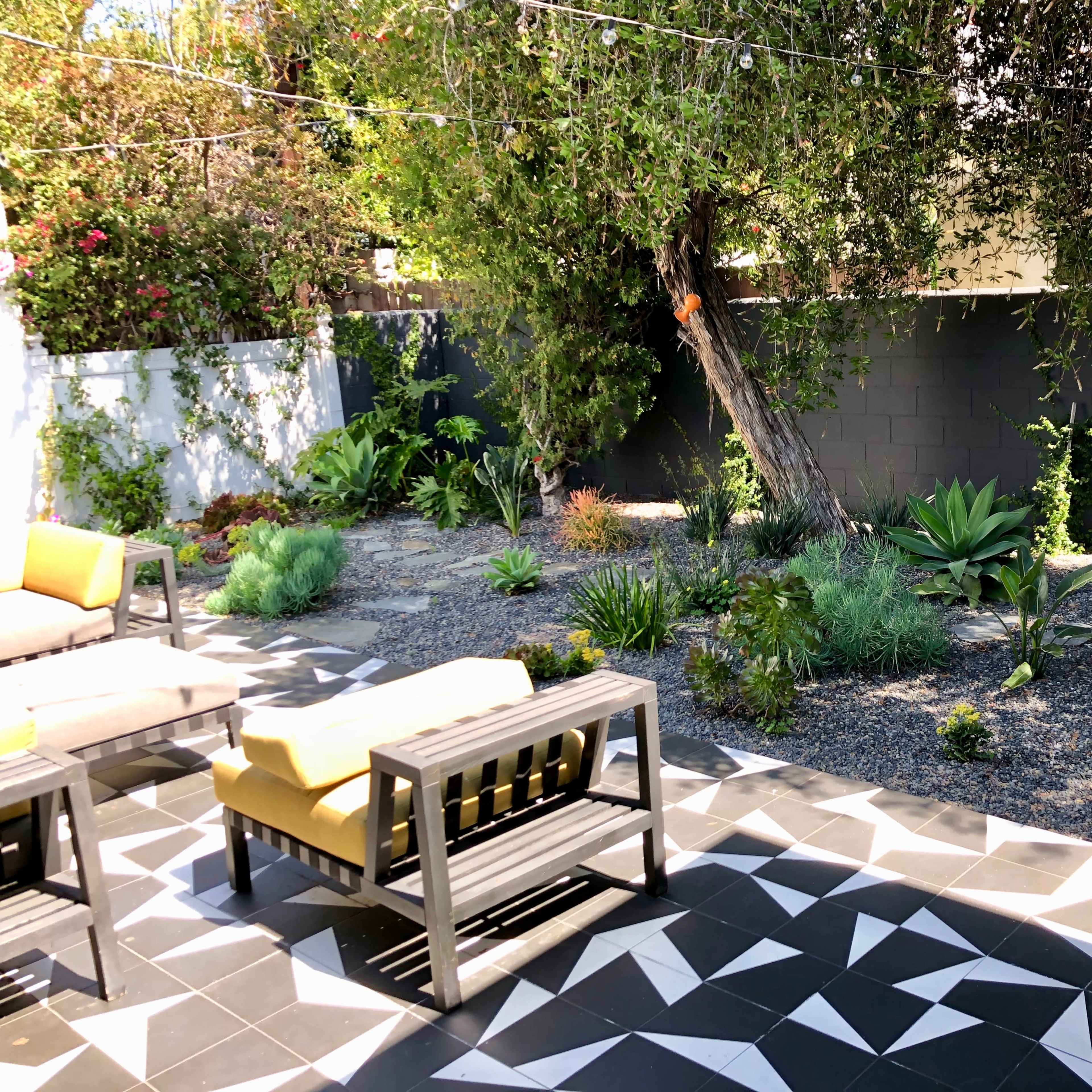 A landscaped garden featuring various plants and gravel, with modern outdoor furniture in a geometric patterned patio area.