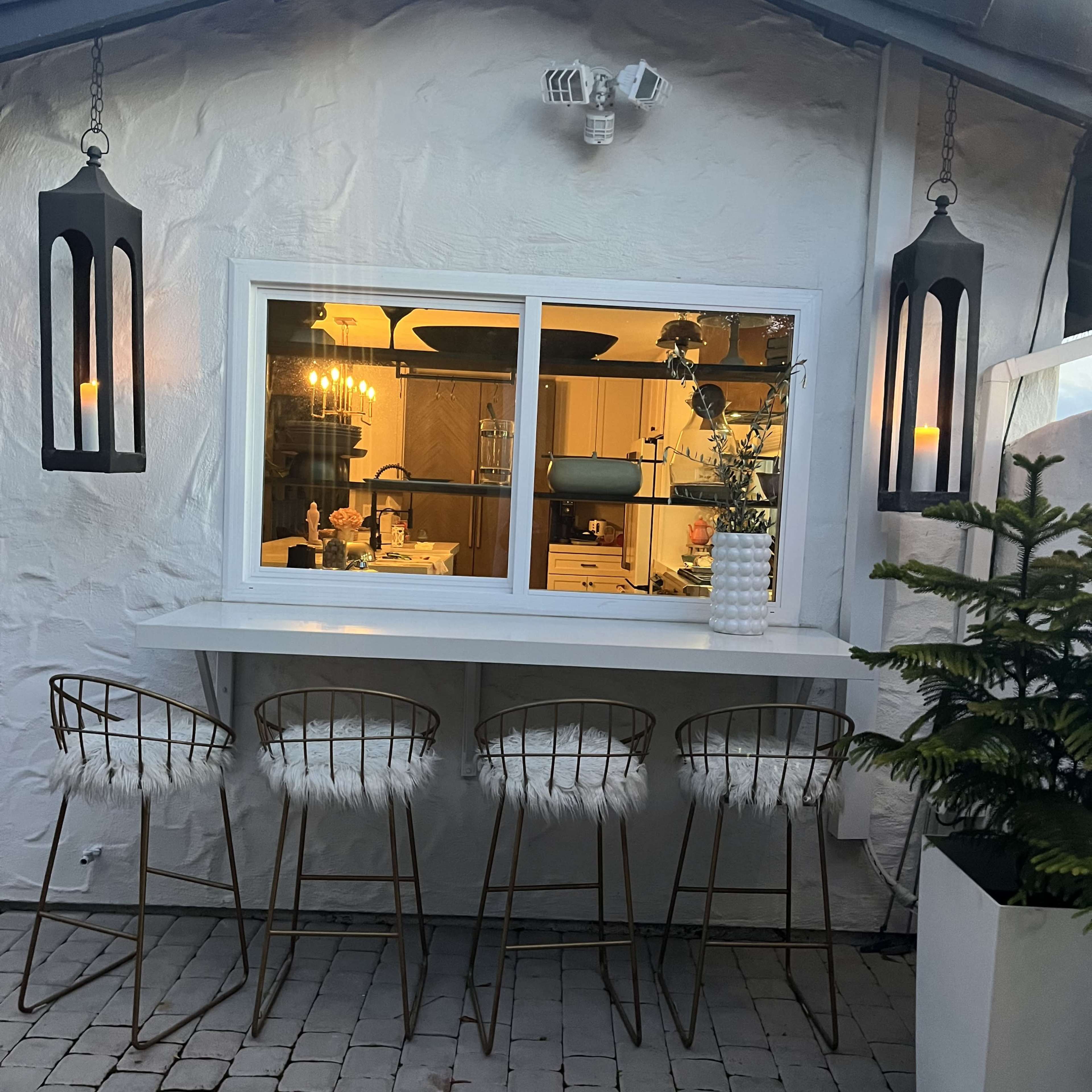 A modern outdoor bar area with four barstools is situated in front of a window that faces a kitchen, framed by two lanterns hanging on either side.