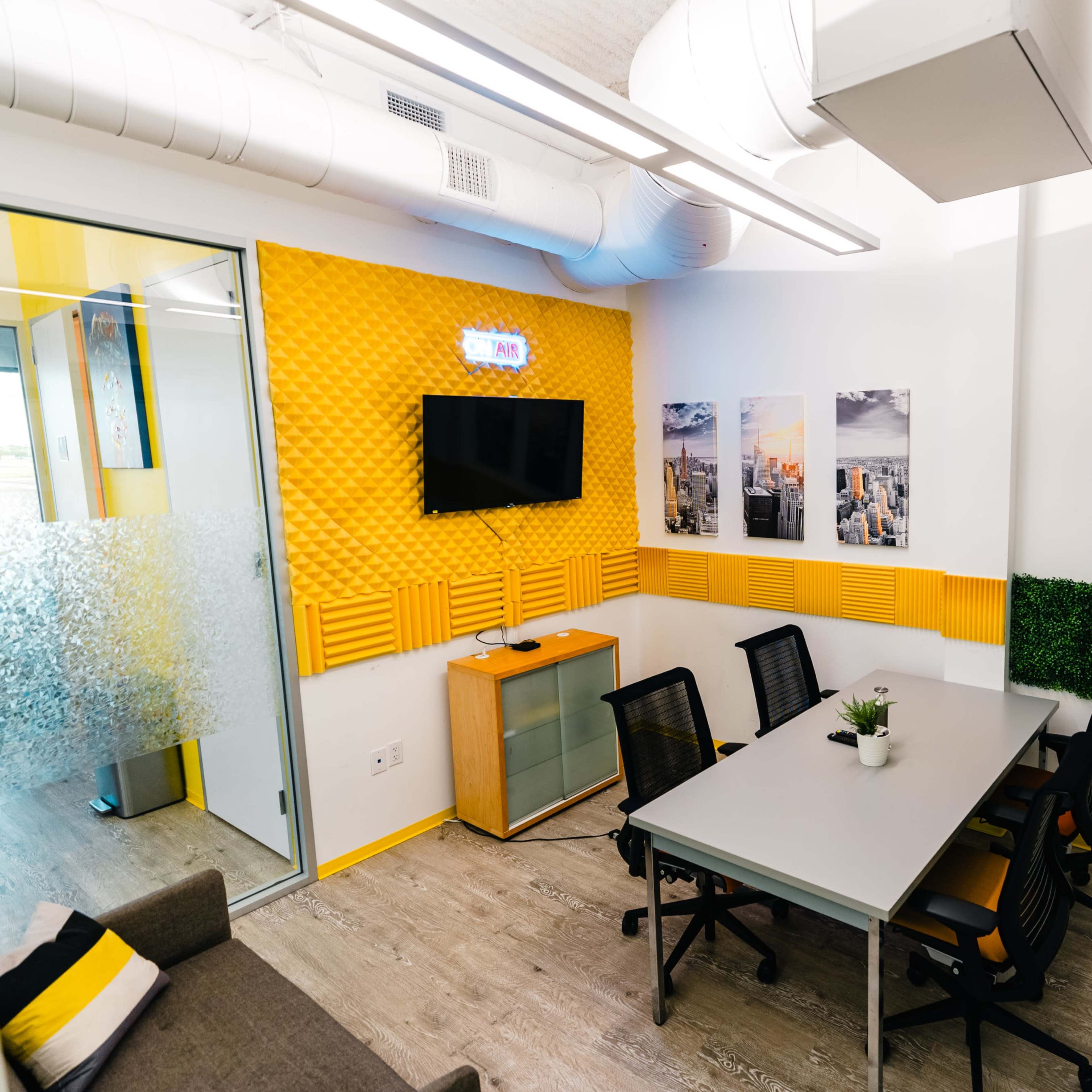 A small meeting room with a glass wall, equipped with a television, a whiteboard, and a table surrounded by chairs, accented by yellow and green decor.