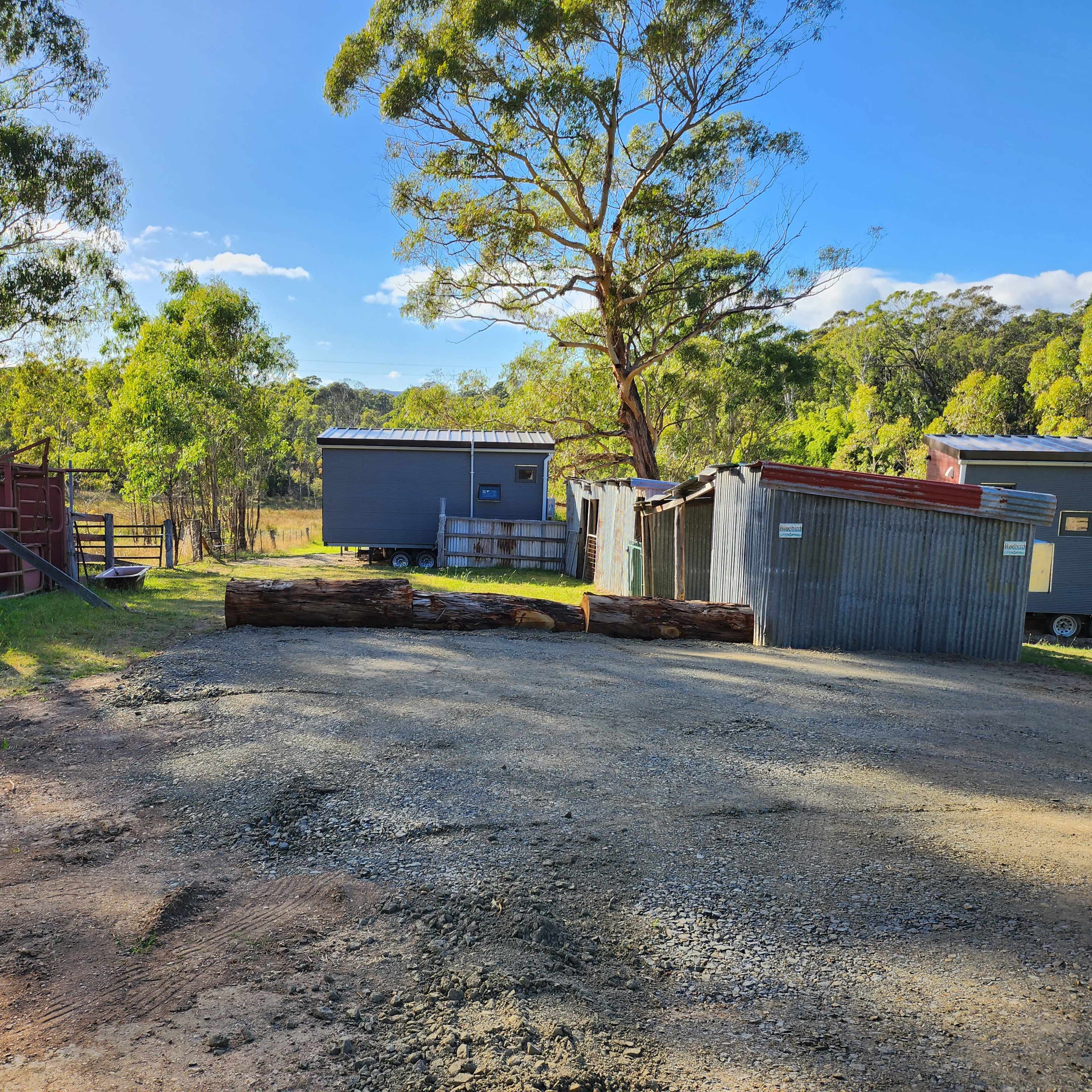 The scene shows a gravel area with several structures, including metal sheds and a caravan, surrounded by trees in a rural setting.