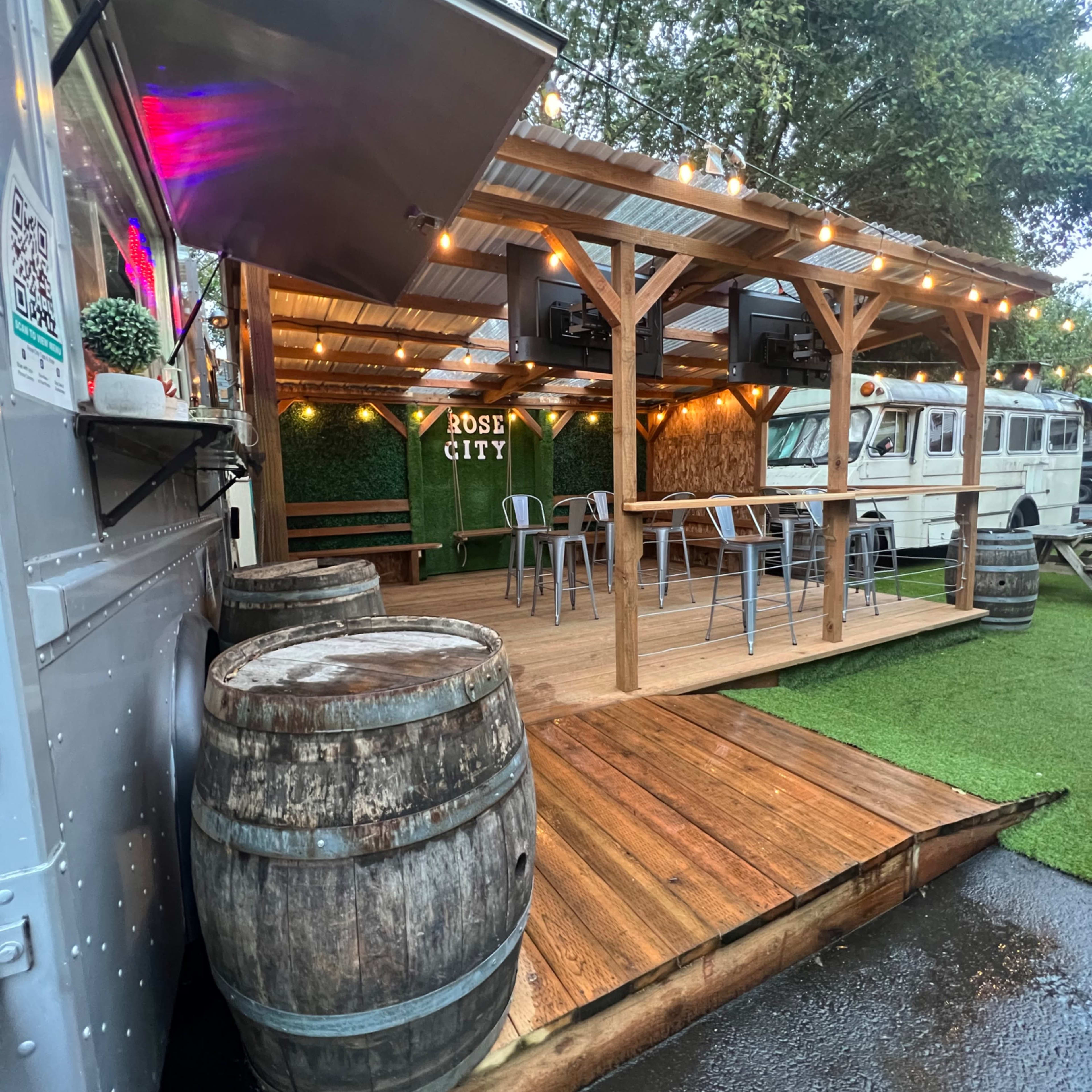 A food truck is positioned next to a wooden deck with floating lights and seating under a covered structure, surrounded by grass and other food trucks.