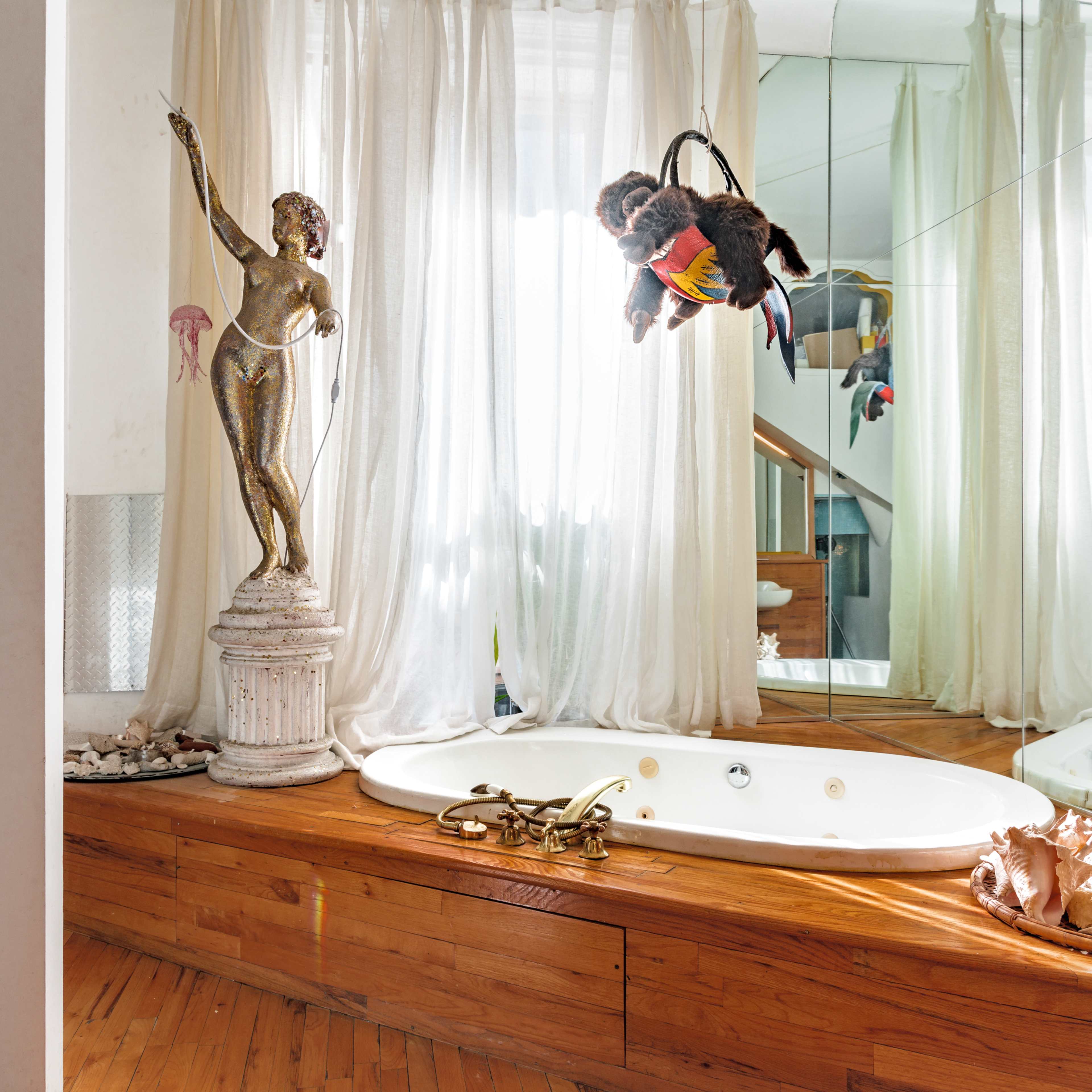 A bathroom with a circular tub surrounded by wooden flooring and a mirror reflecting a statue and various decor items.