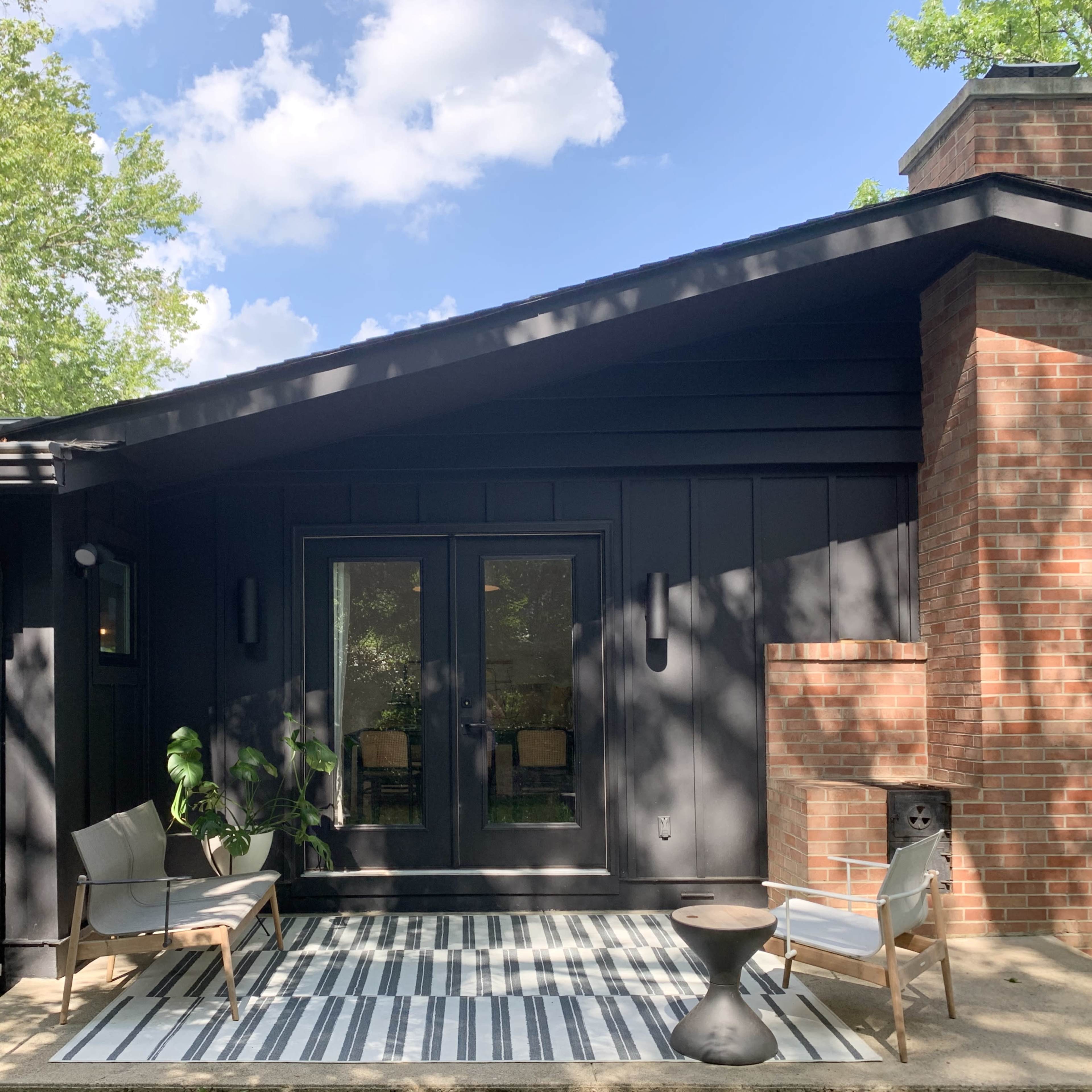 A modern black wood-panelled house with large glass doors opening onto a small patio featuring two chairs and a striped rug.
