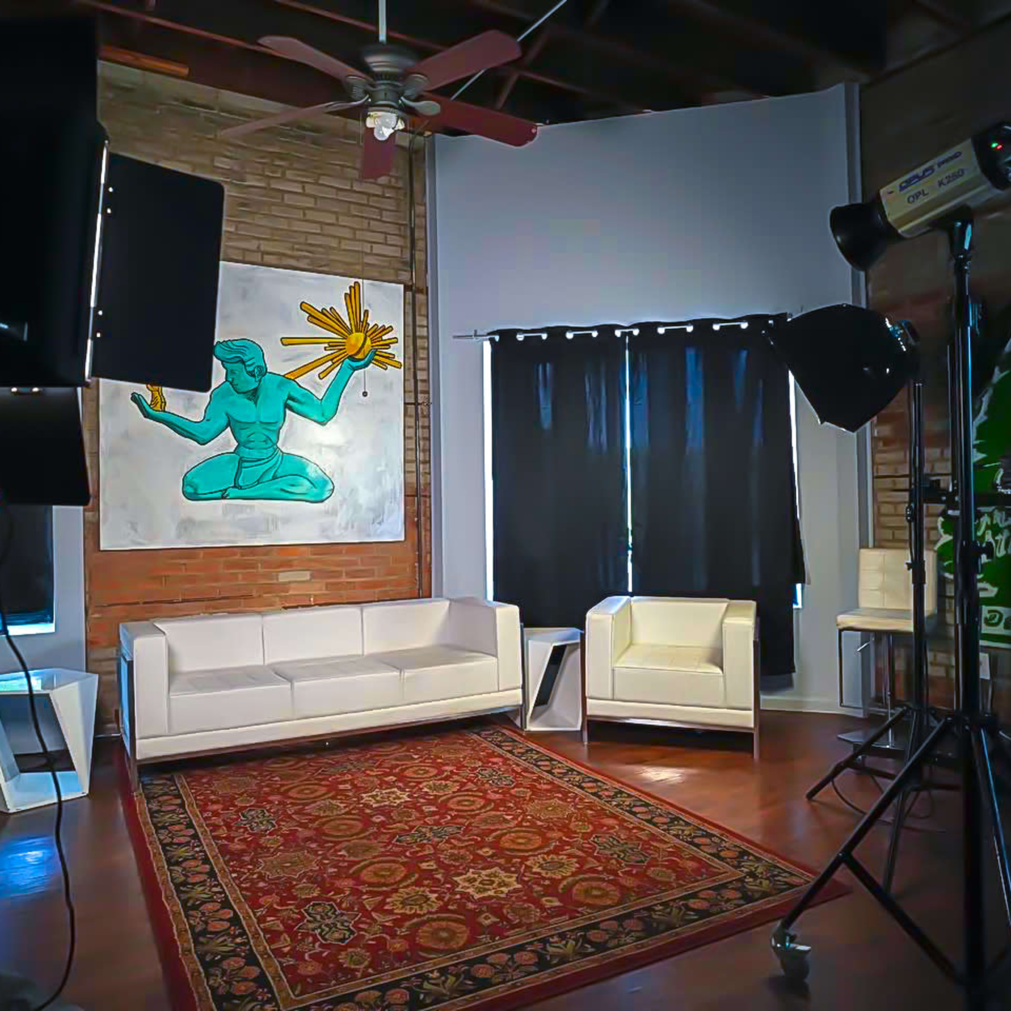 A well-lit studio space featuring a white sofa, a white armchair, a colorful wall painting, and photography lighting equipment.