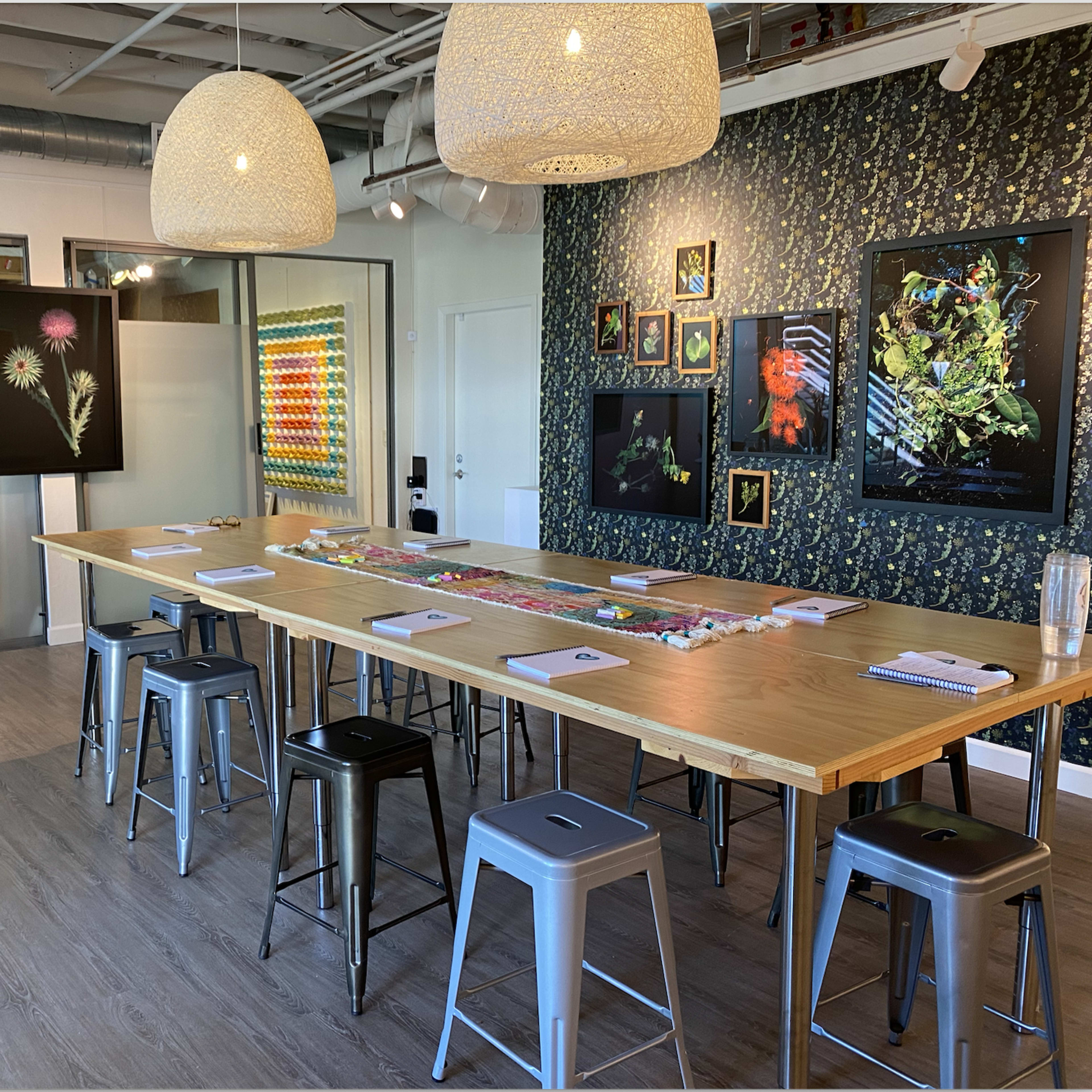 A modern dining or meeting space with a long wooden table surrounded by metal stools, decorated walls, and framed artworks displayed.