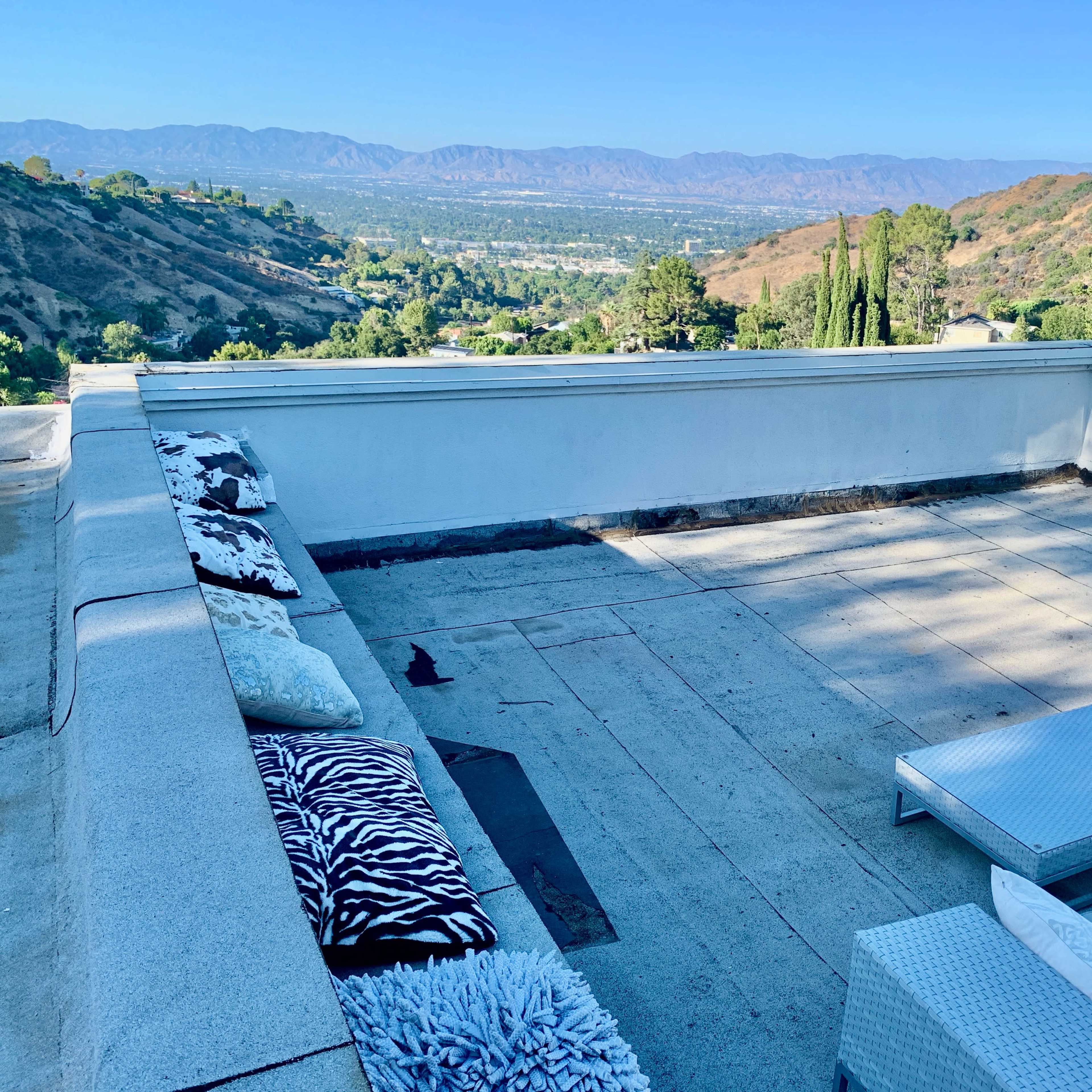 A rooftop terrace with several loungers and cushions, overlooking a landscape of hills and mountains under a clear blue sky.
