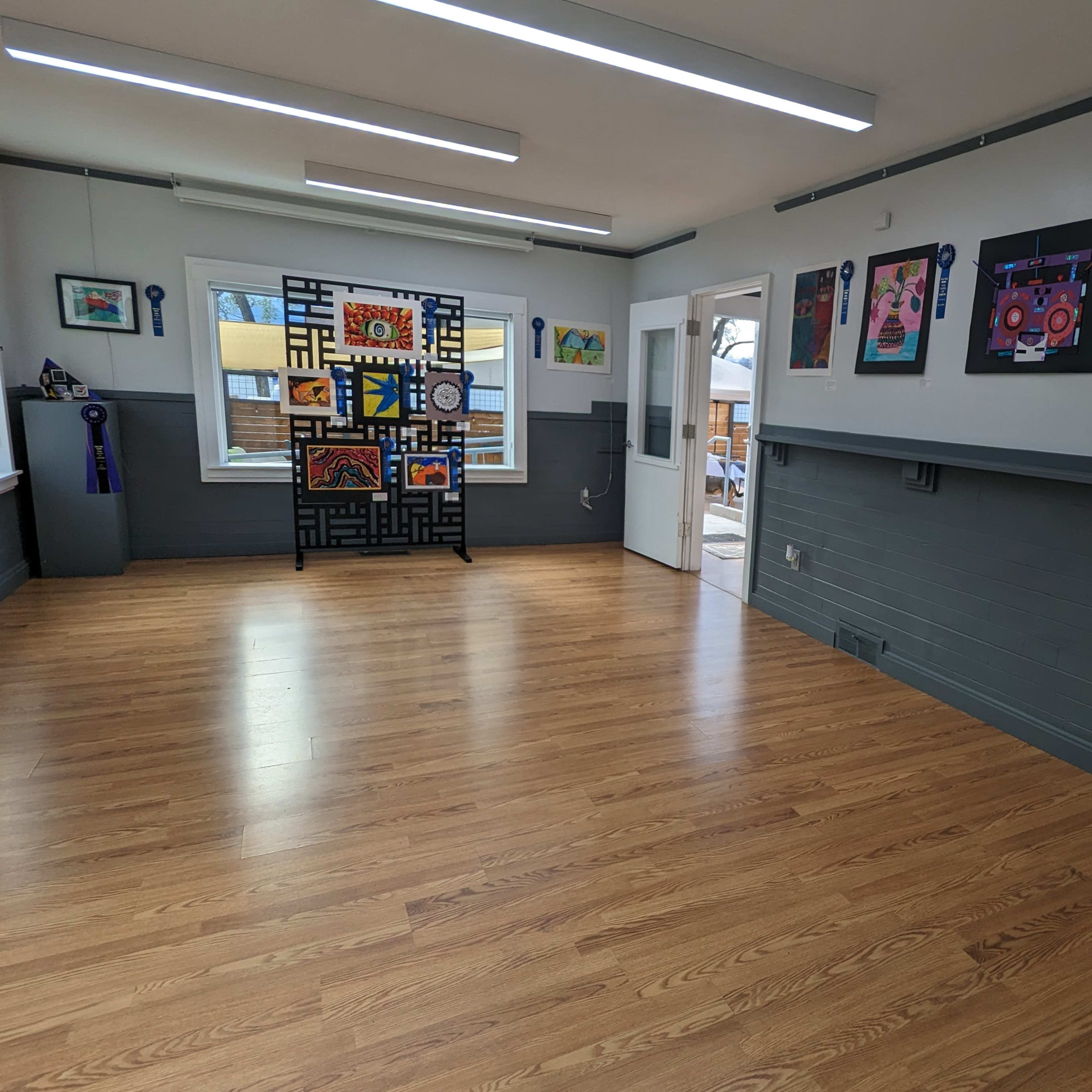 An empty room with wooden flooring, featuring colorful artwork displayed on the walls and a partition with abstract designs.