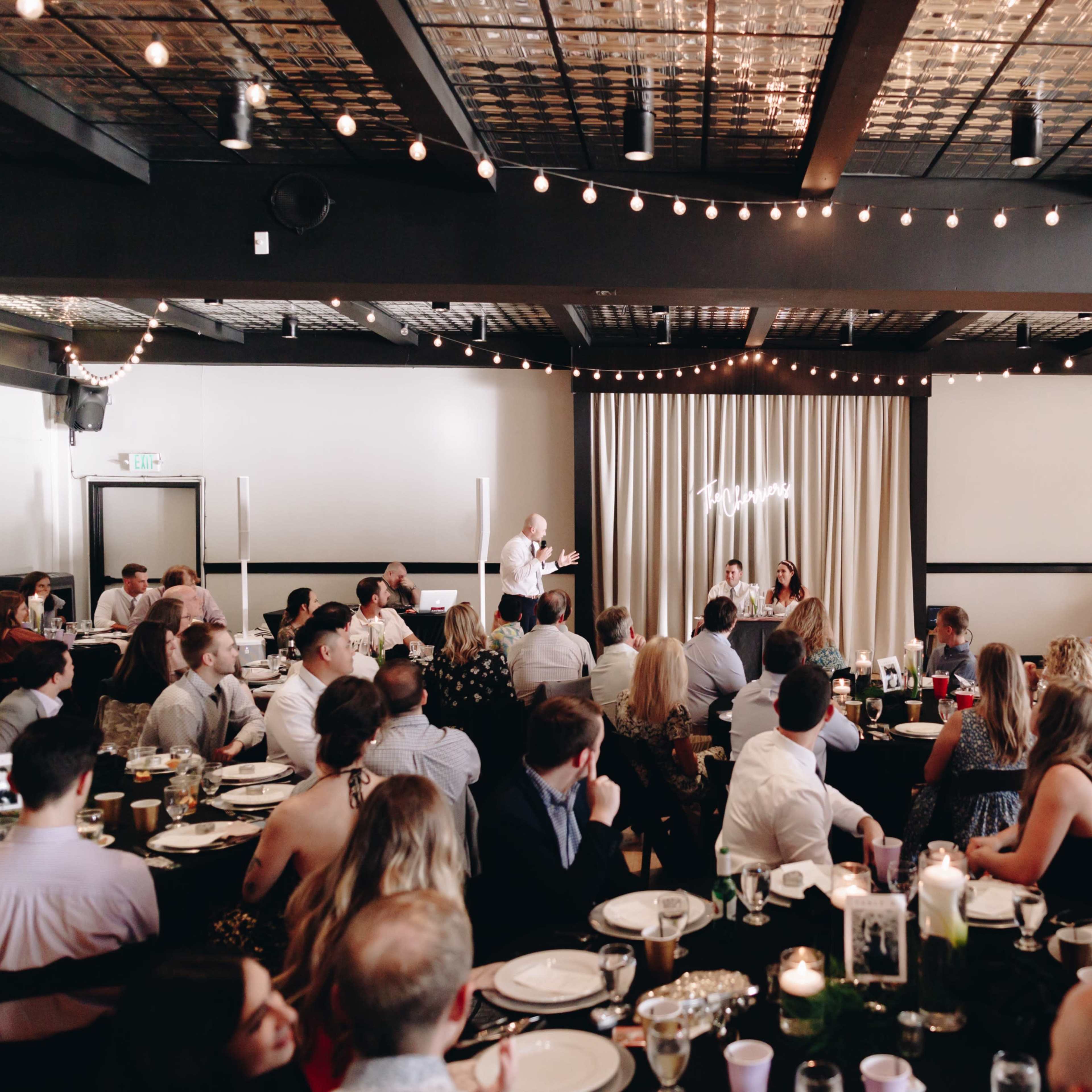 A crowd of guests sits at elegantly set tables in a decorated reception hall, while a speaker addresses the audience near a softly lit backdrop.
