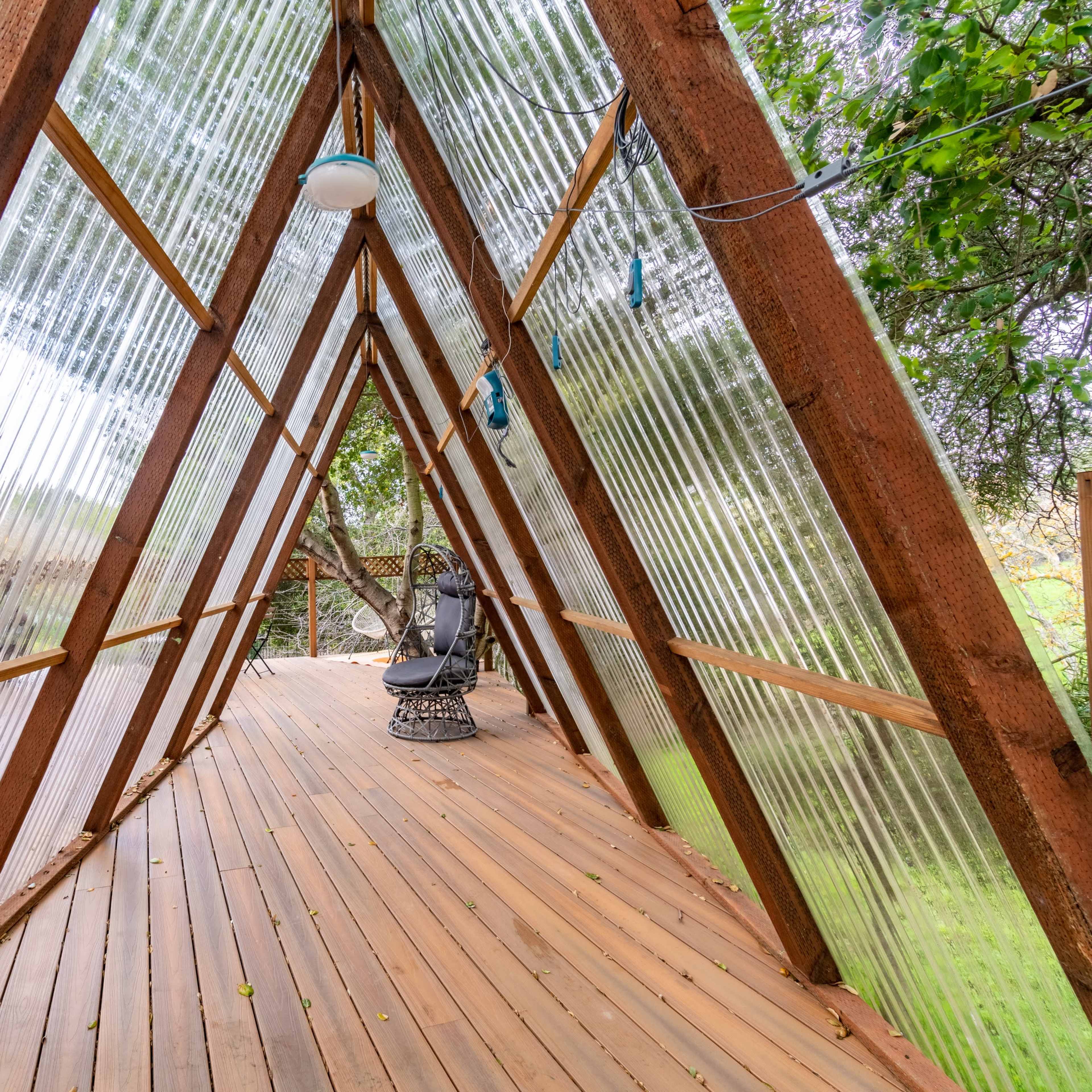 A wooden deck in an A-frame structure with transparent walls, surrounded by greenery.