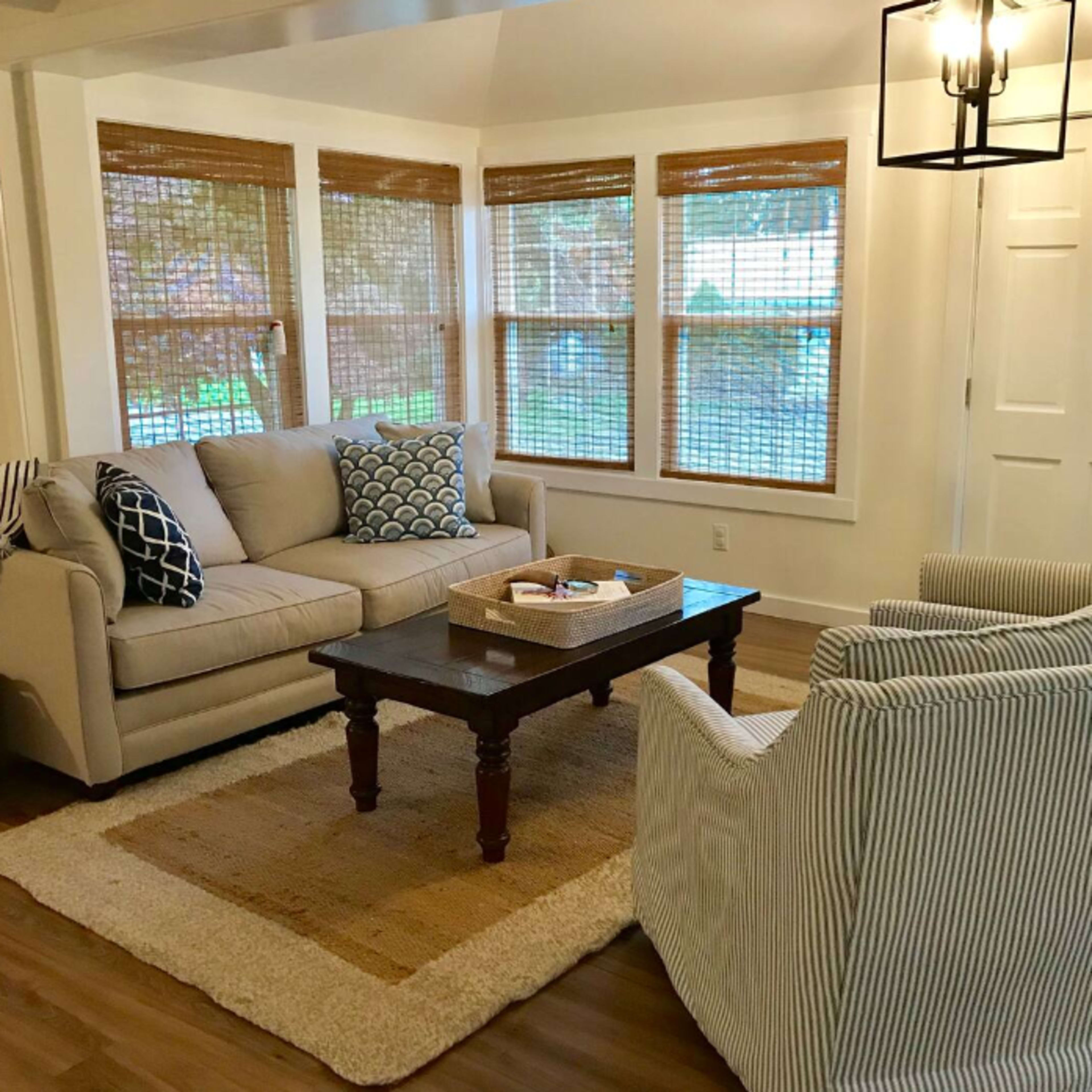 A living room features a beige sofa and two striped armchairs arranged around a wooden coffee table, with large windows adorned with bamboo shades and a front door nearby.