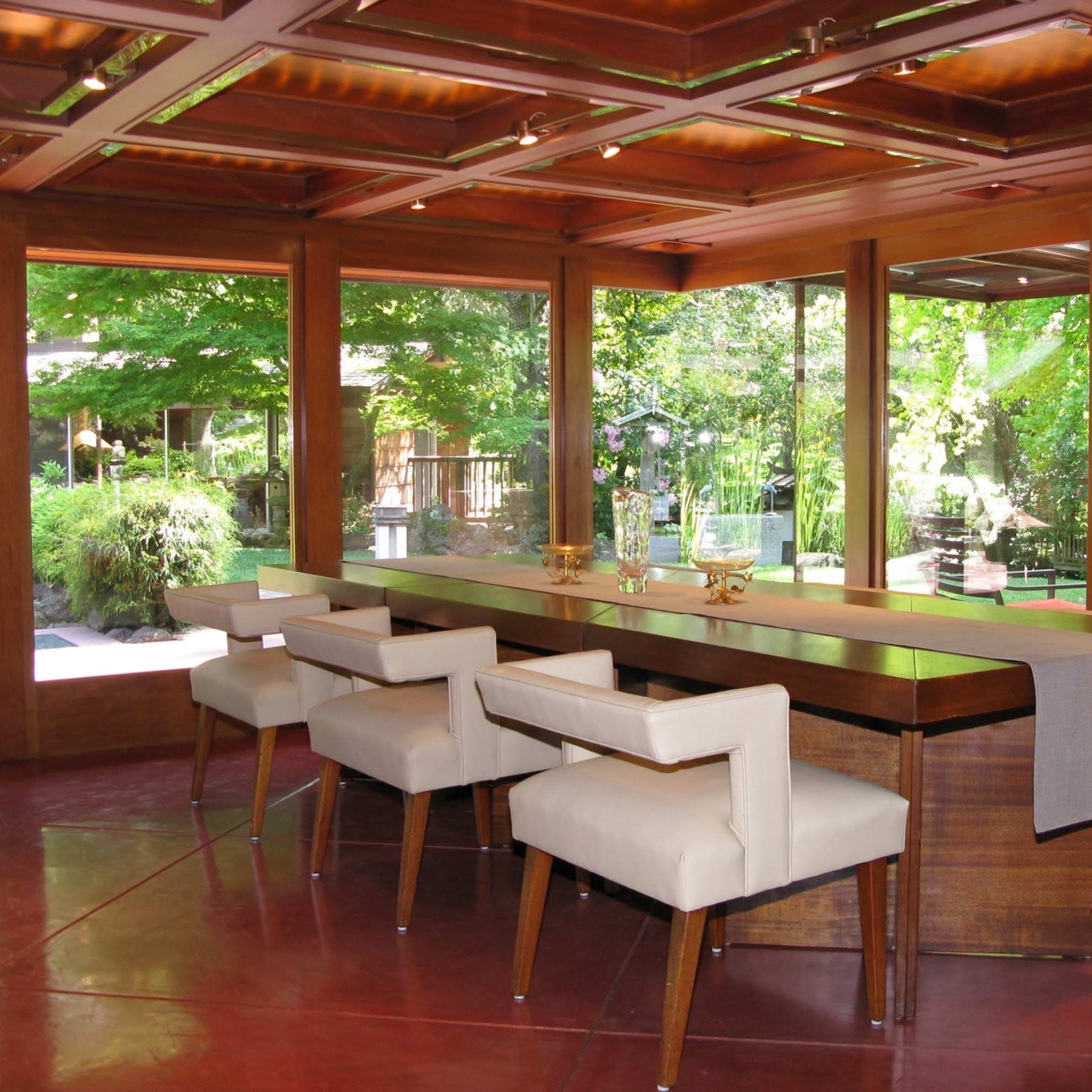 A contemporary dining area with a long wooden table surrounded by four upholstered chairs, overlooking a lush outdoor garden through large windows.