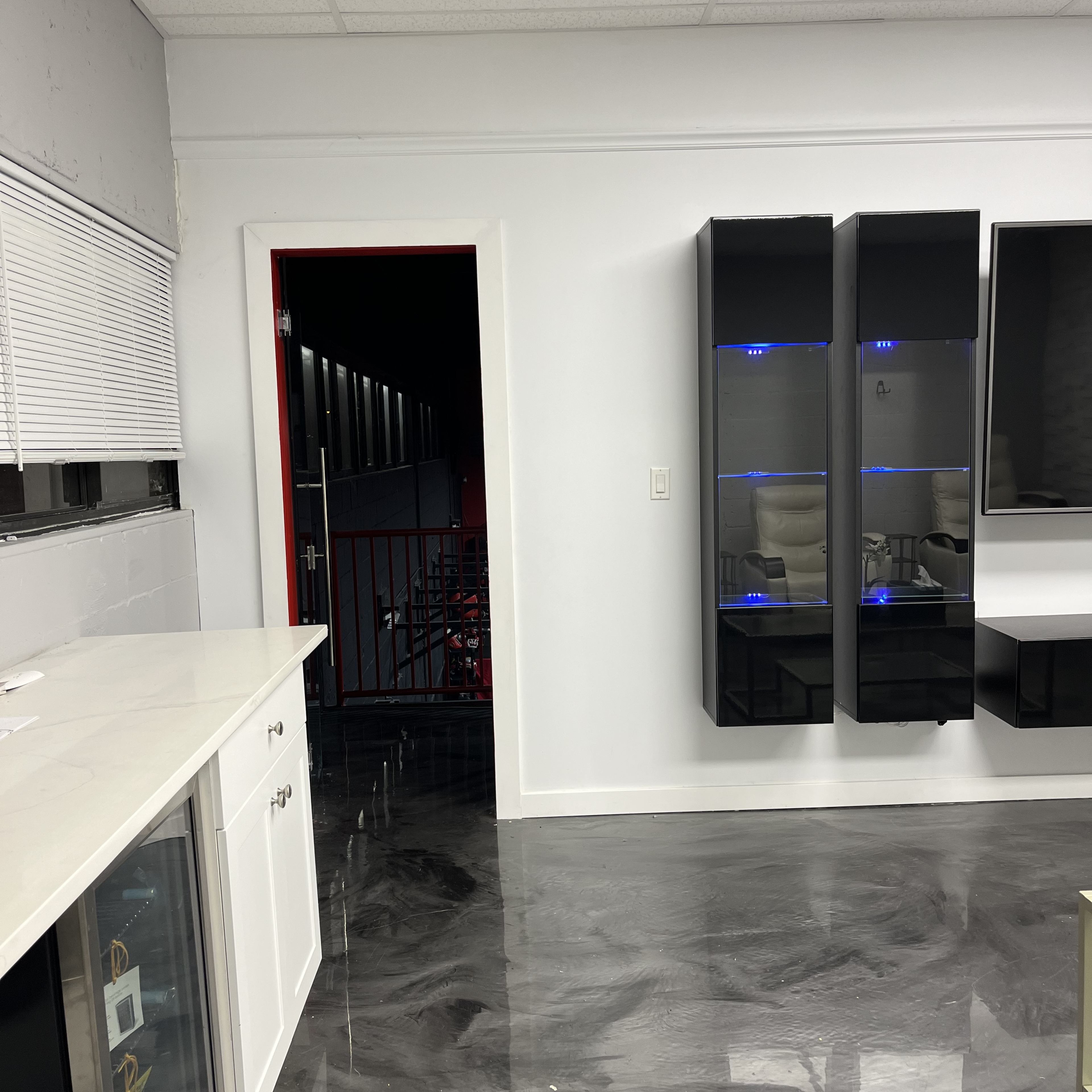 A modern room with a white wall, dark cabinets, a countertop with a sink, and a door leading to a dimly lit area.