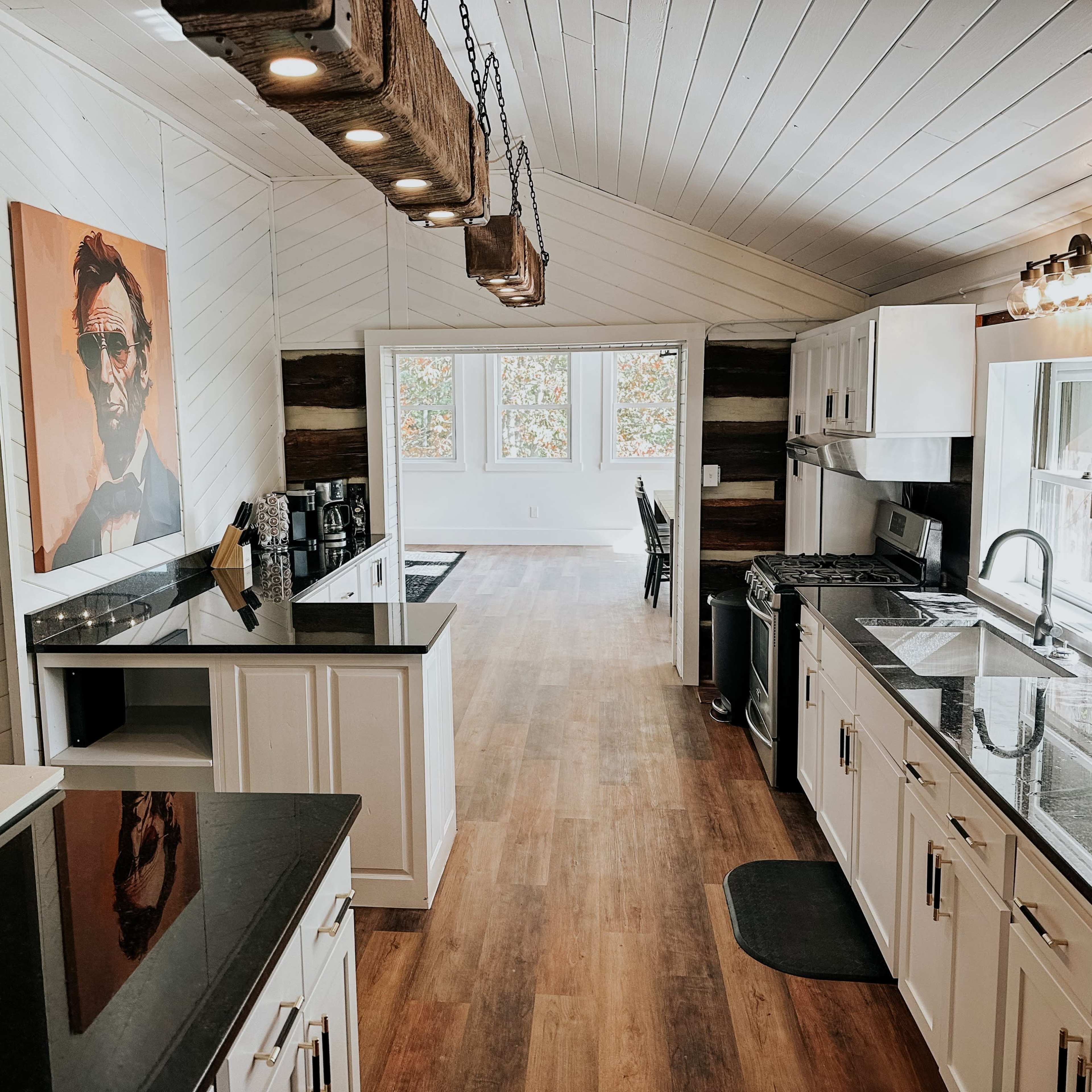 A modern kitchen with white cabinets, dark countertops, and a large window, featuring an art piece on the wall.