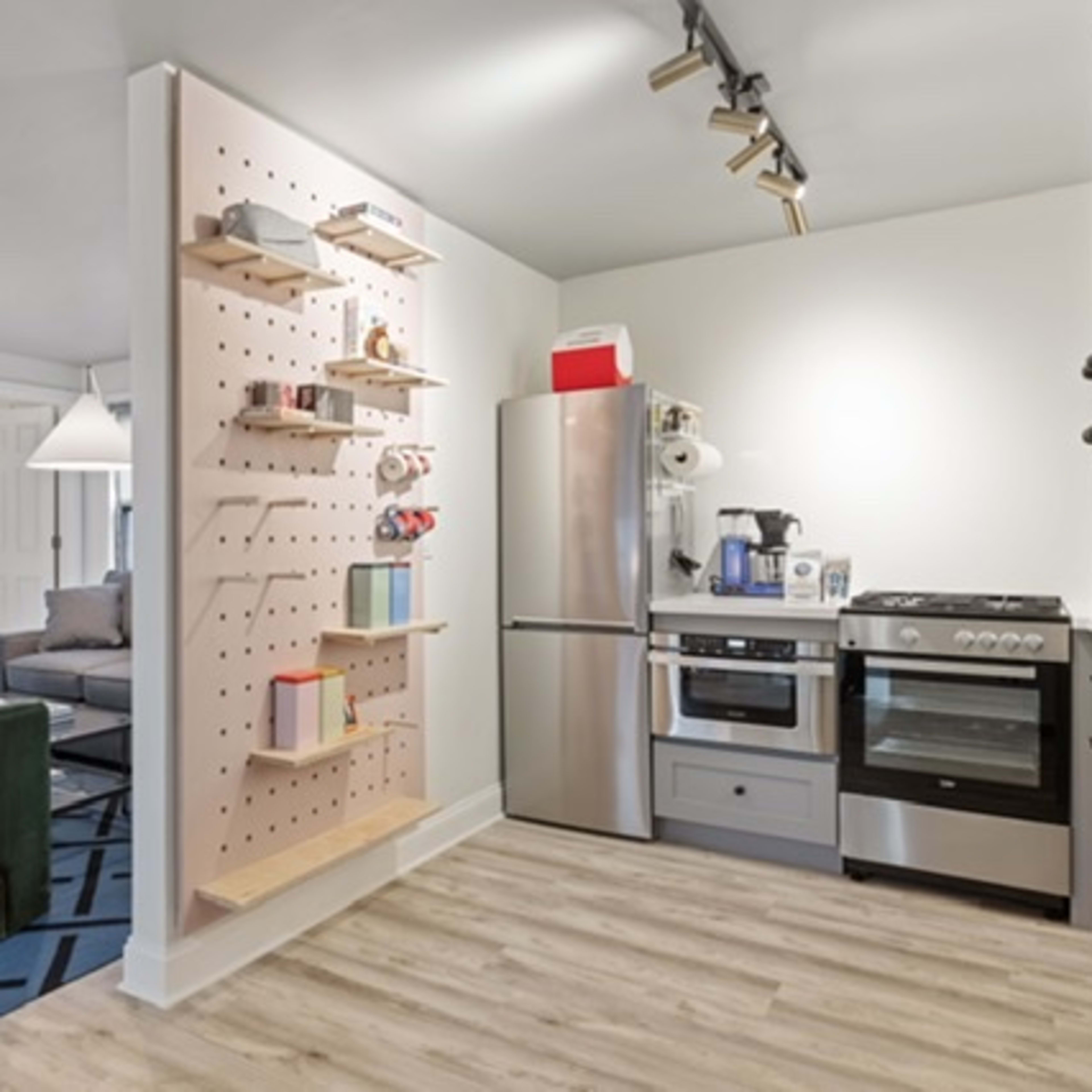 A modern kitchen and living area featuring a pegboard shelf, stainless steel appliances, and two green armchairs on a patterned rug.
