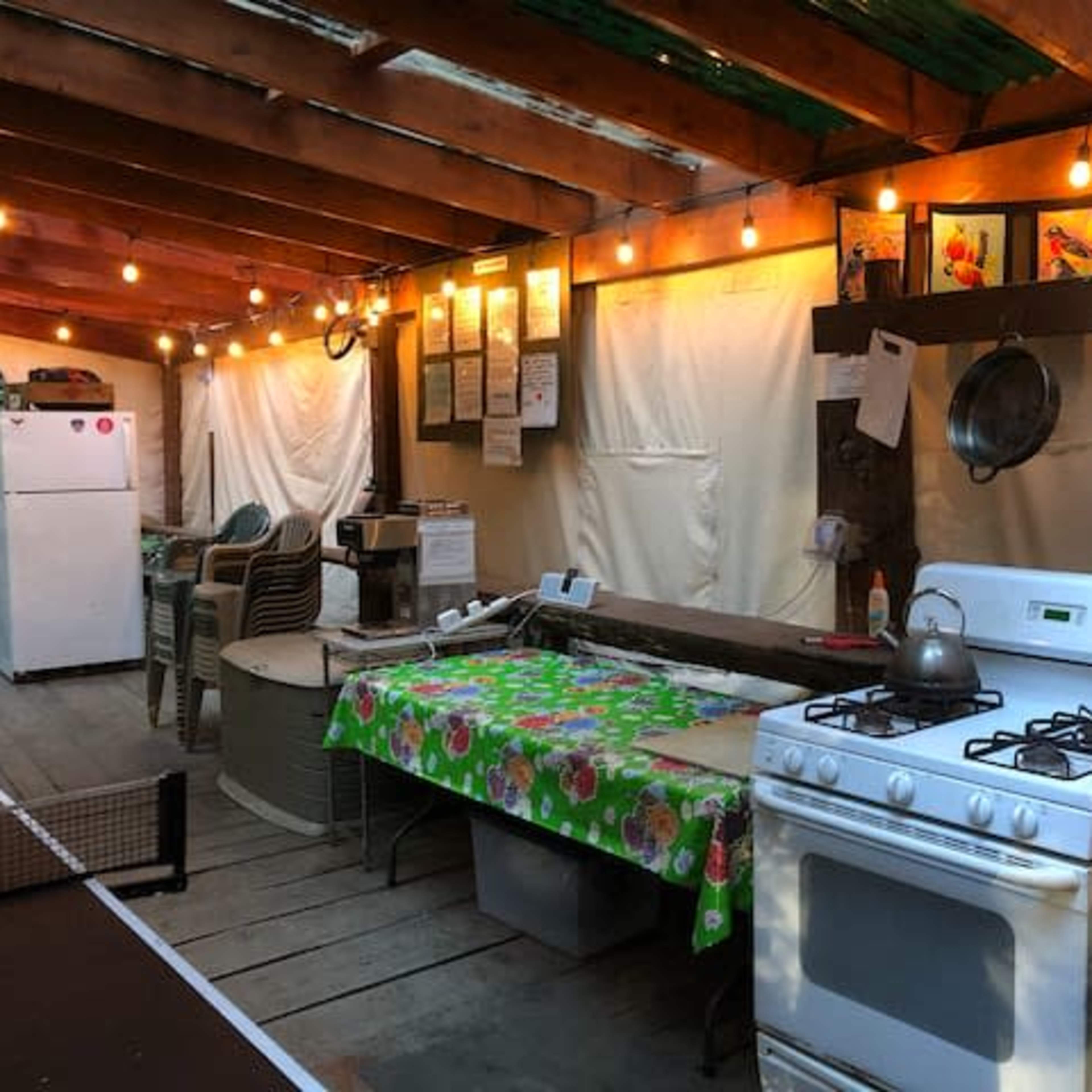 A rustic outdoor kitchen featuring a gas stove, a table covered with a floral tablecloth, and various hanging pots and light strings overhead.