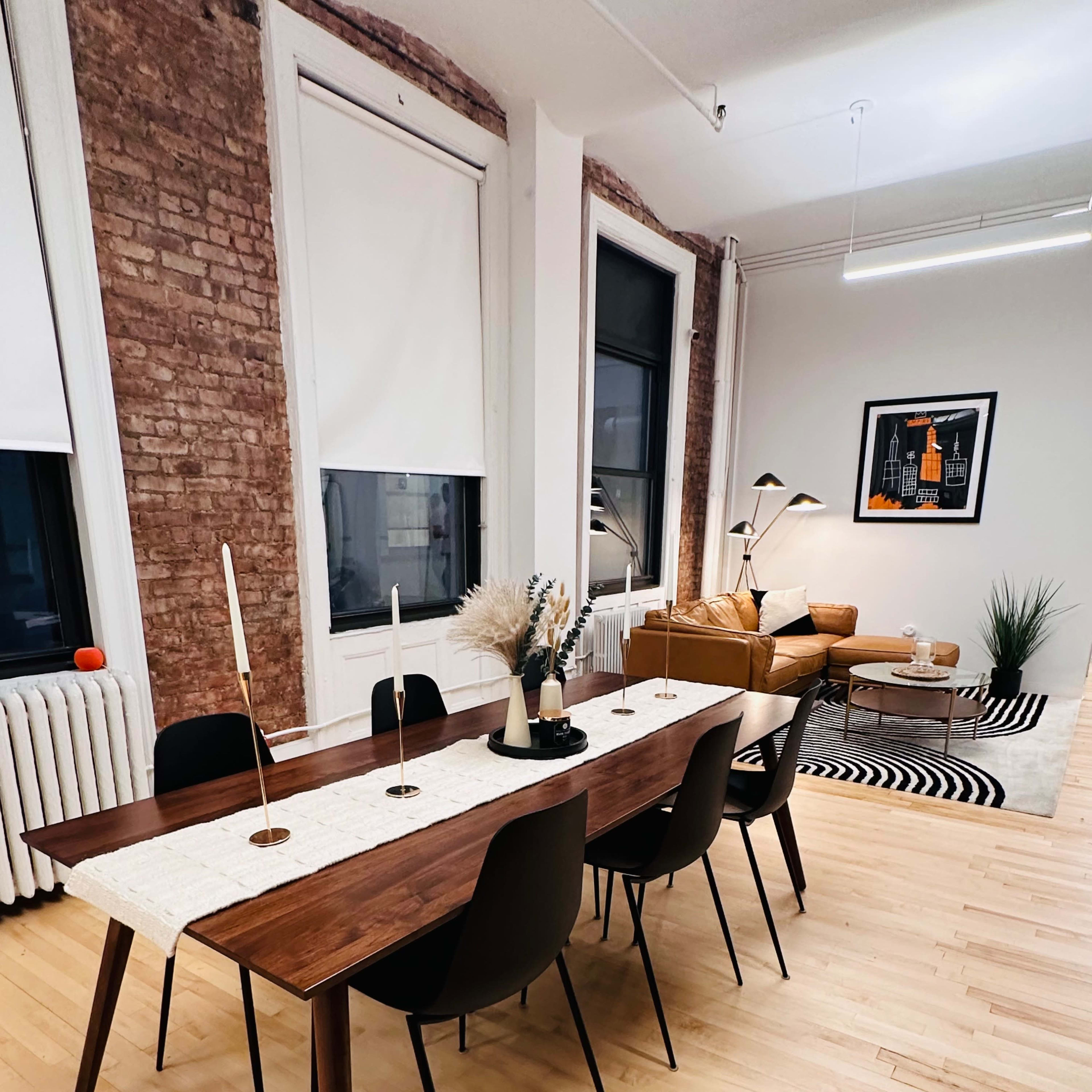 A long wooden dining table with black chairs and candles is set against a backdrop of exposed brick walls and large windows in a modern living space.