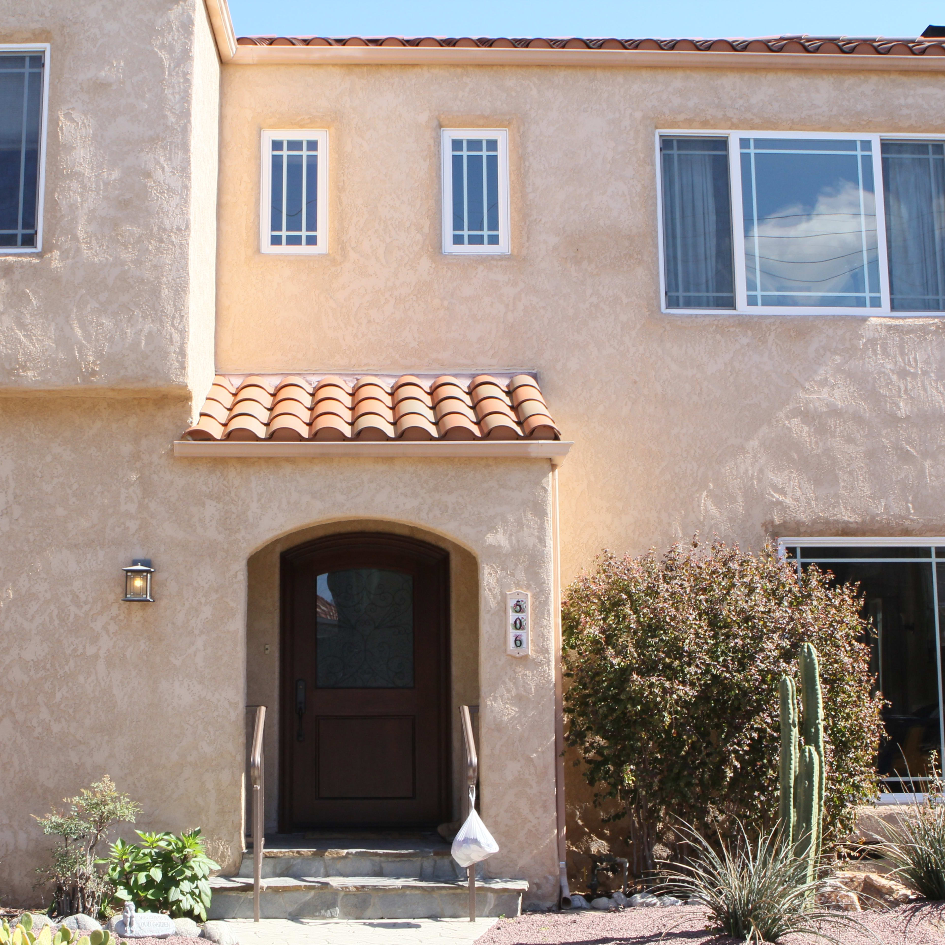 A two-story stucco house with a tiled roof, featuring multiple windows, a wooden front door, and desert landscaping with cacti.