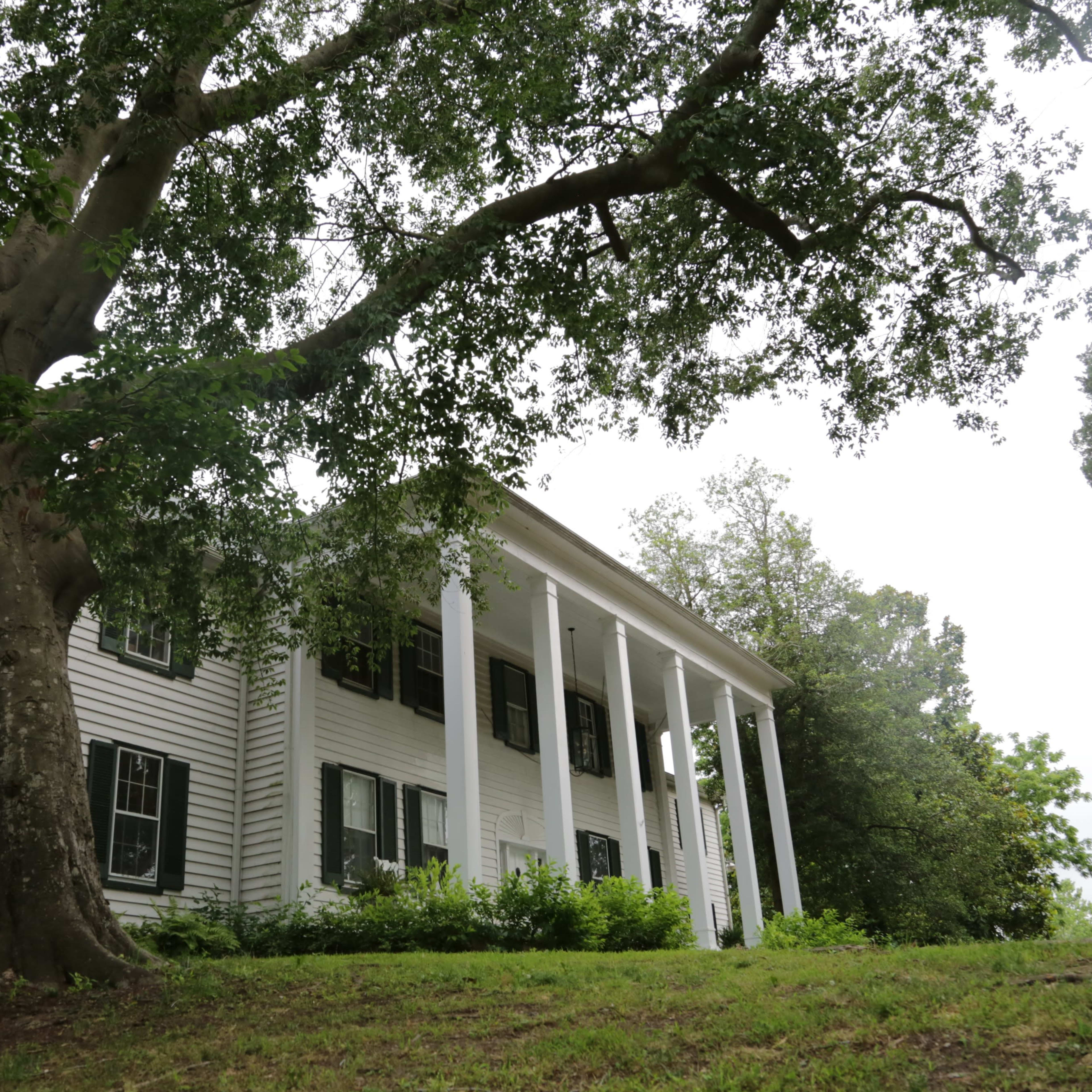 A large white house with six columns stands on a gentle slope beside a prominent tree.