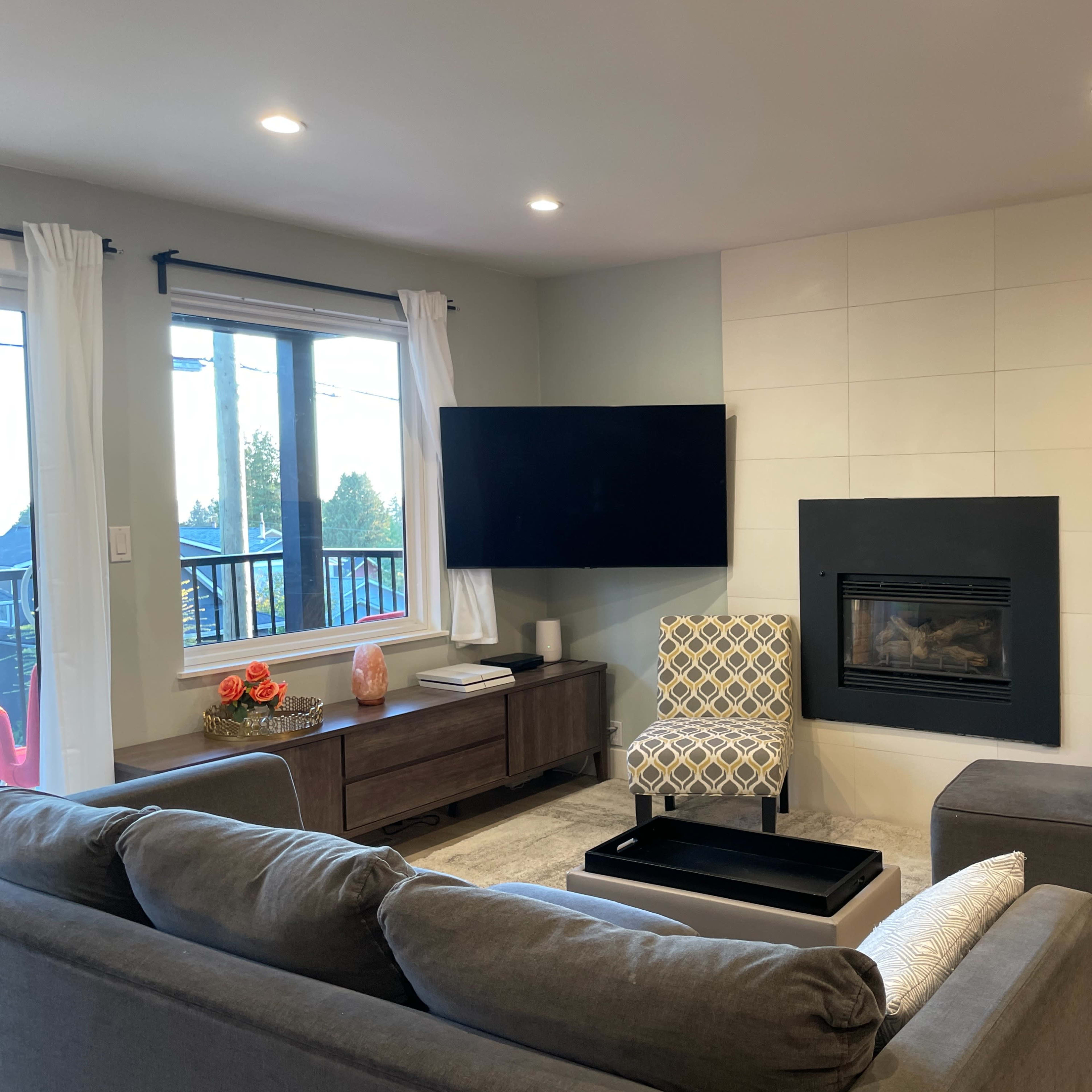 A living room with a television mounted on the wall, a fireplace, a gray sofa set, and large windows allowing natural light to enter.