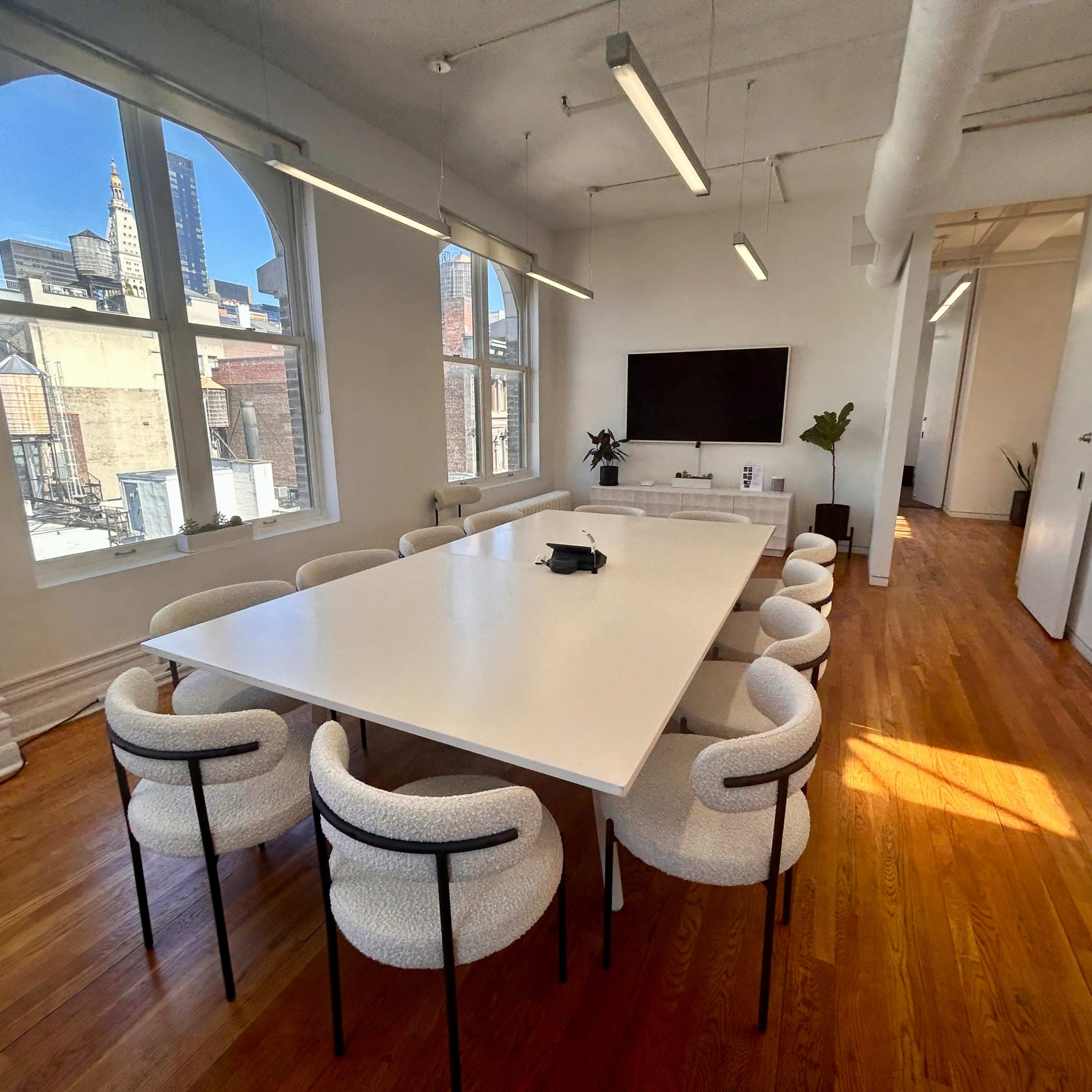 A modern conference room features a large white table surrounded by upholstered chairs, with windows revealing a city skyline.