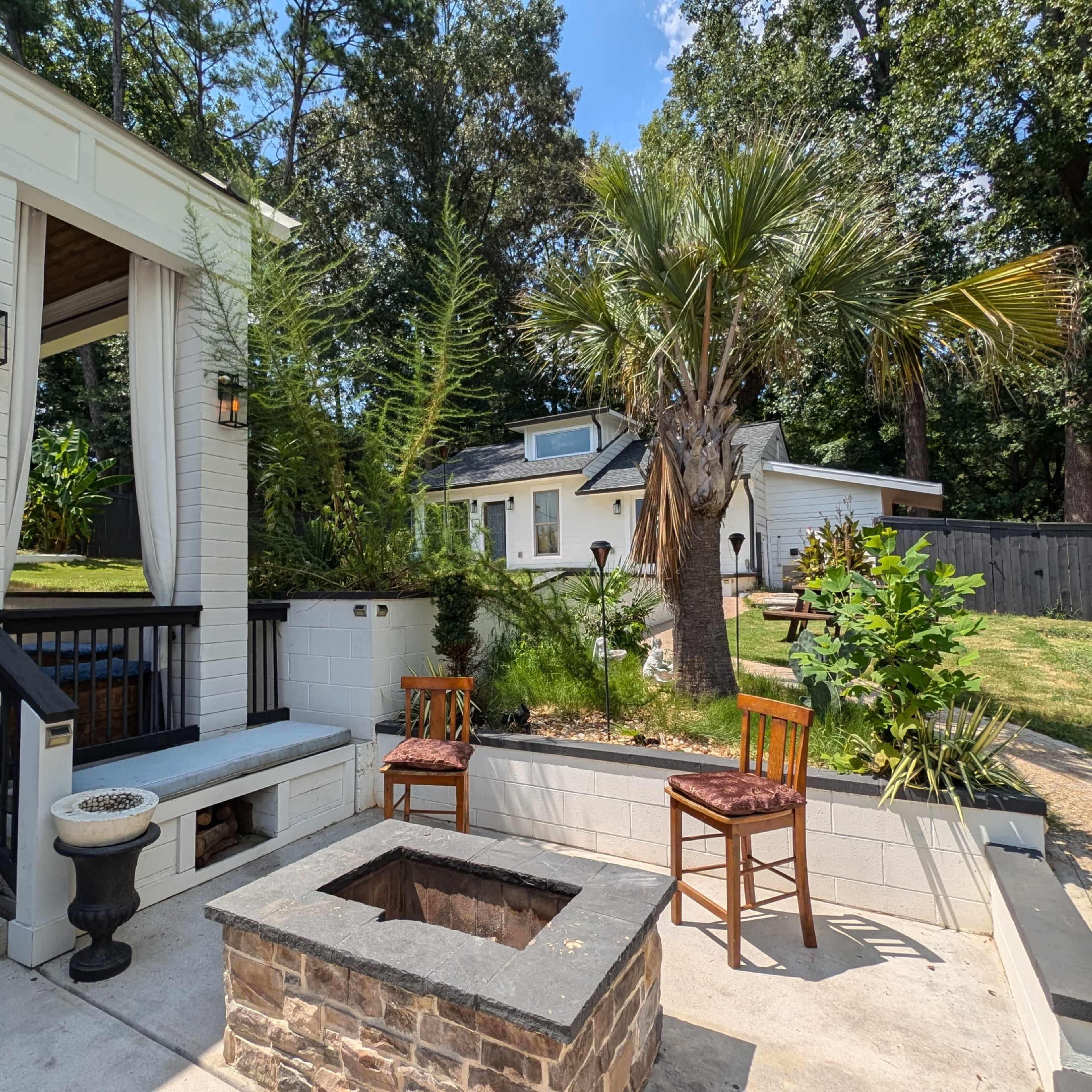 A backyard patio area with a fire pit surrounded by two chairs, a small white house in the background, and palm trees framing the scene.