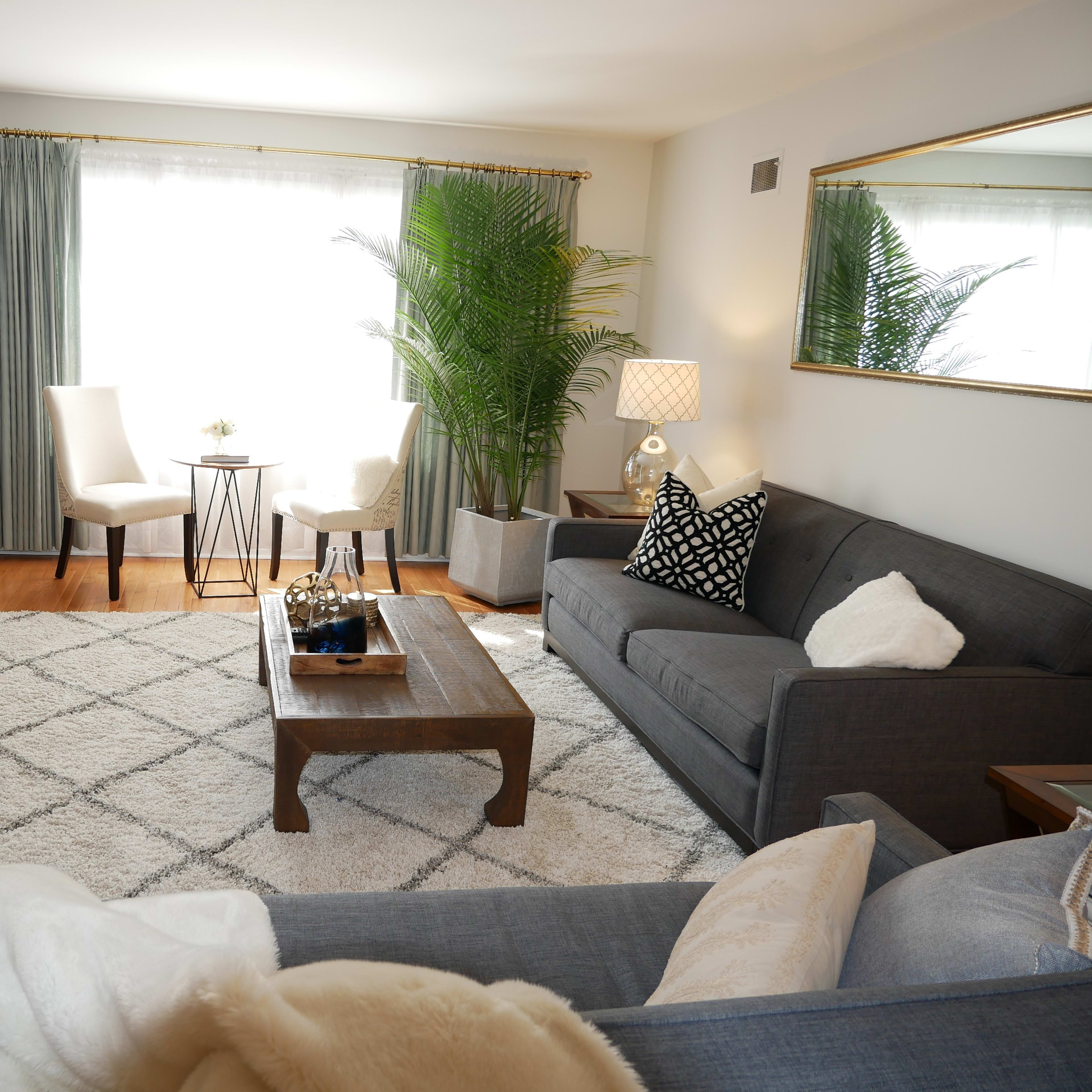 A cozy living room with a gray sofa, a wooden coffee table, two chairs, and large windows allowing natural light to enter.