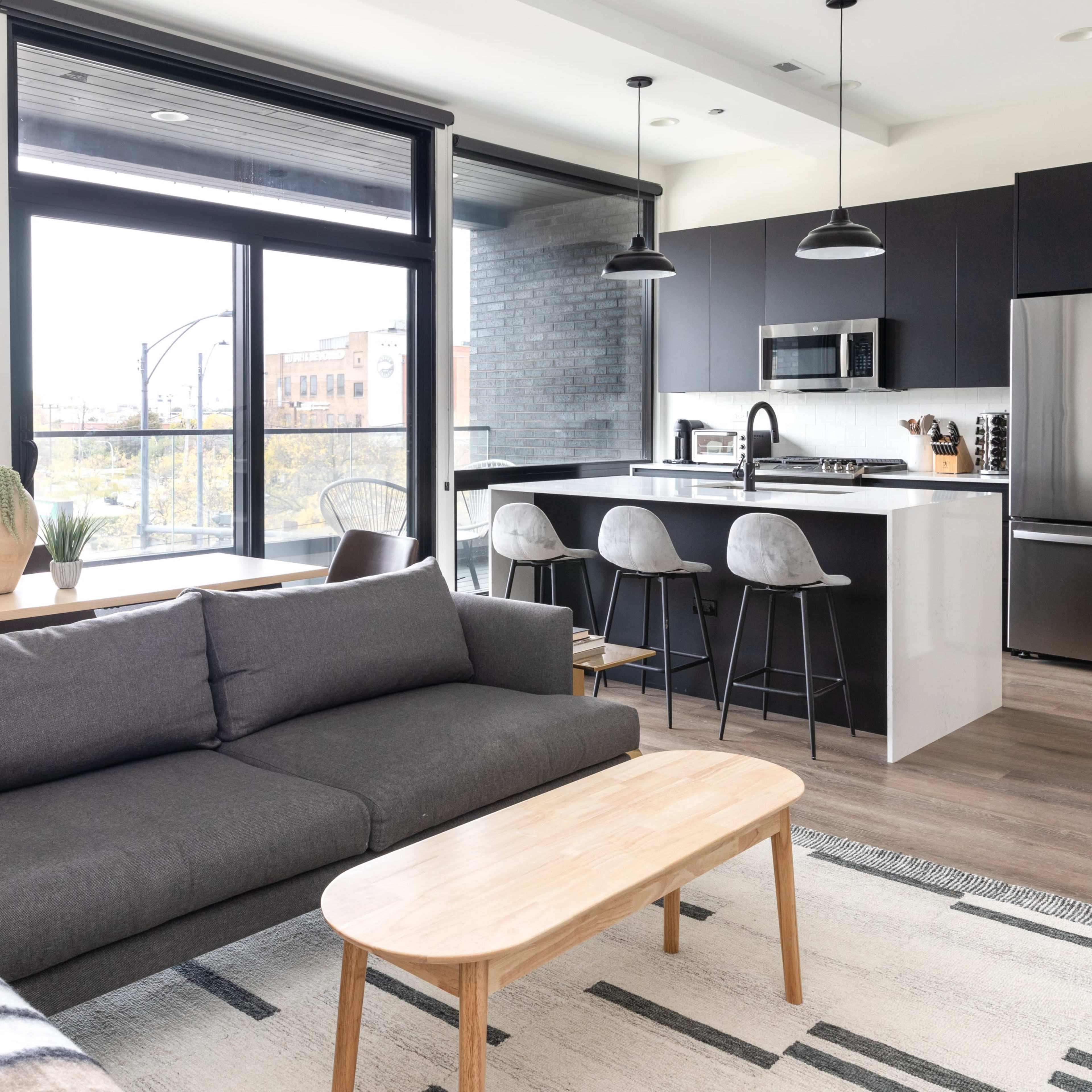 A modern living space featuring a gray sofa, a light wood coffee table, and a kitchen area with dark cabinetry and bar stools.