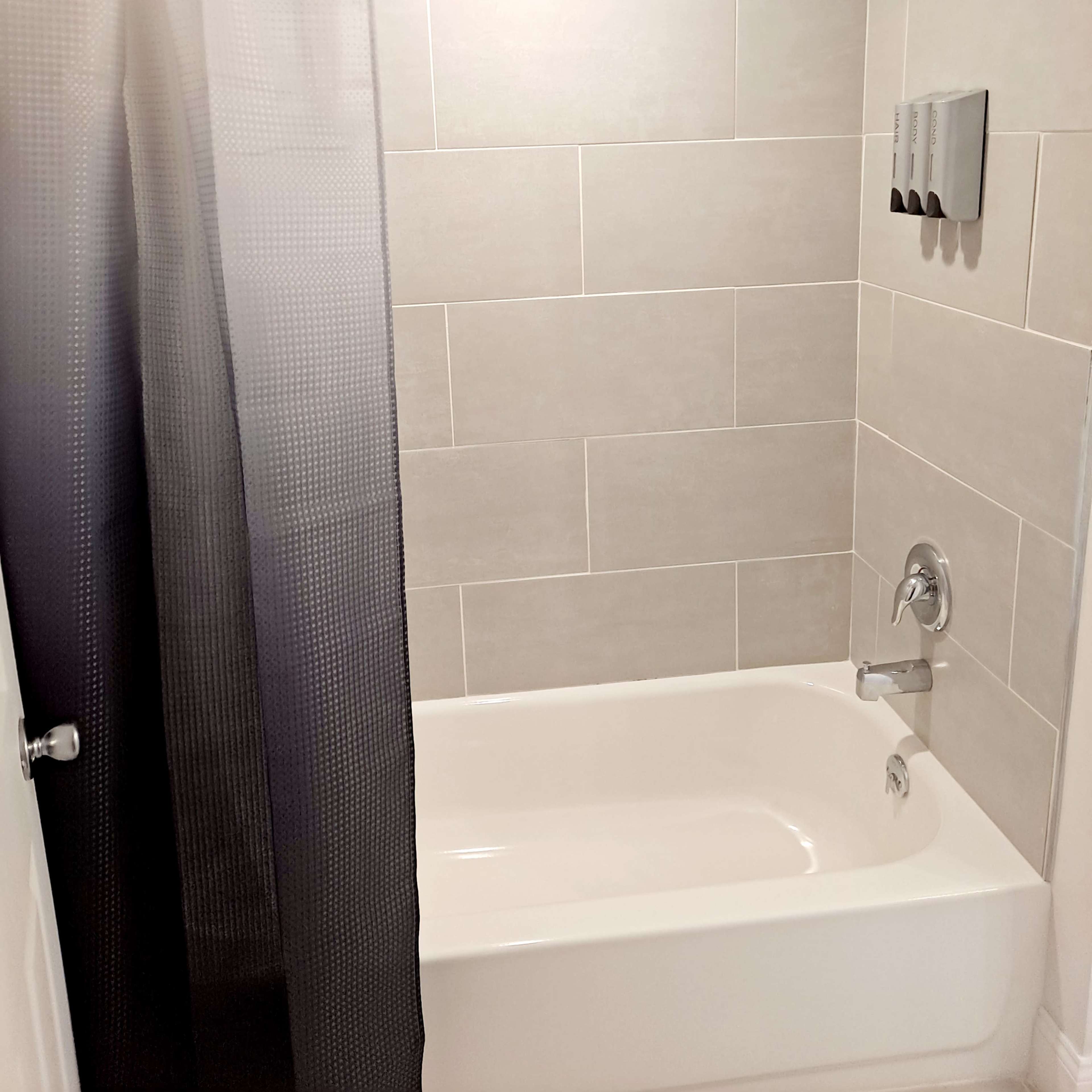 A bathroom featuring a shower area with a gray tiled wall, a white bathtub, a showerhead, and a shower curtain partially covering the space.