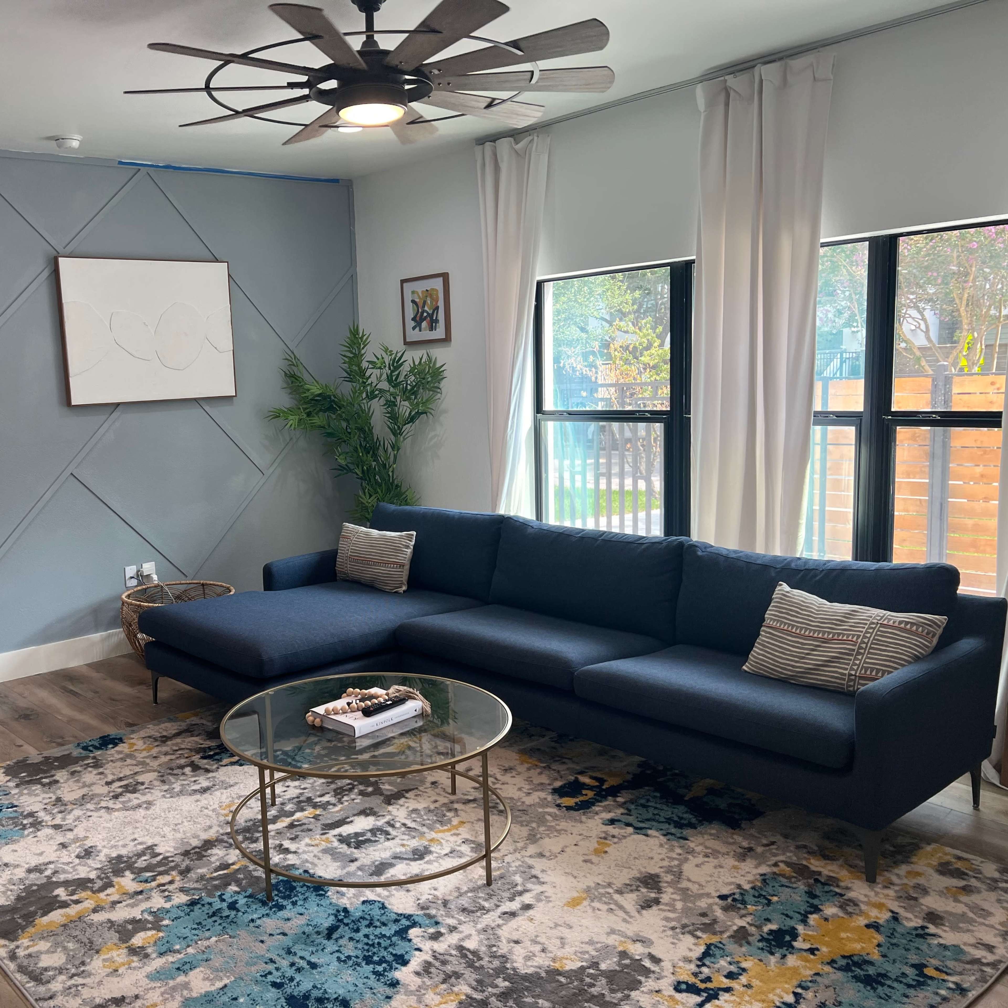 A modern living room featuring a blue sectional sofa, a round glass coffee table, and large windows with white curtains.
