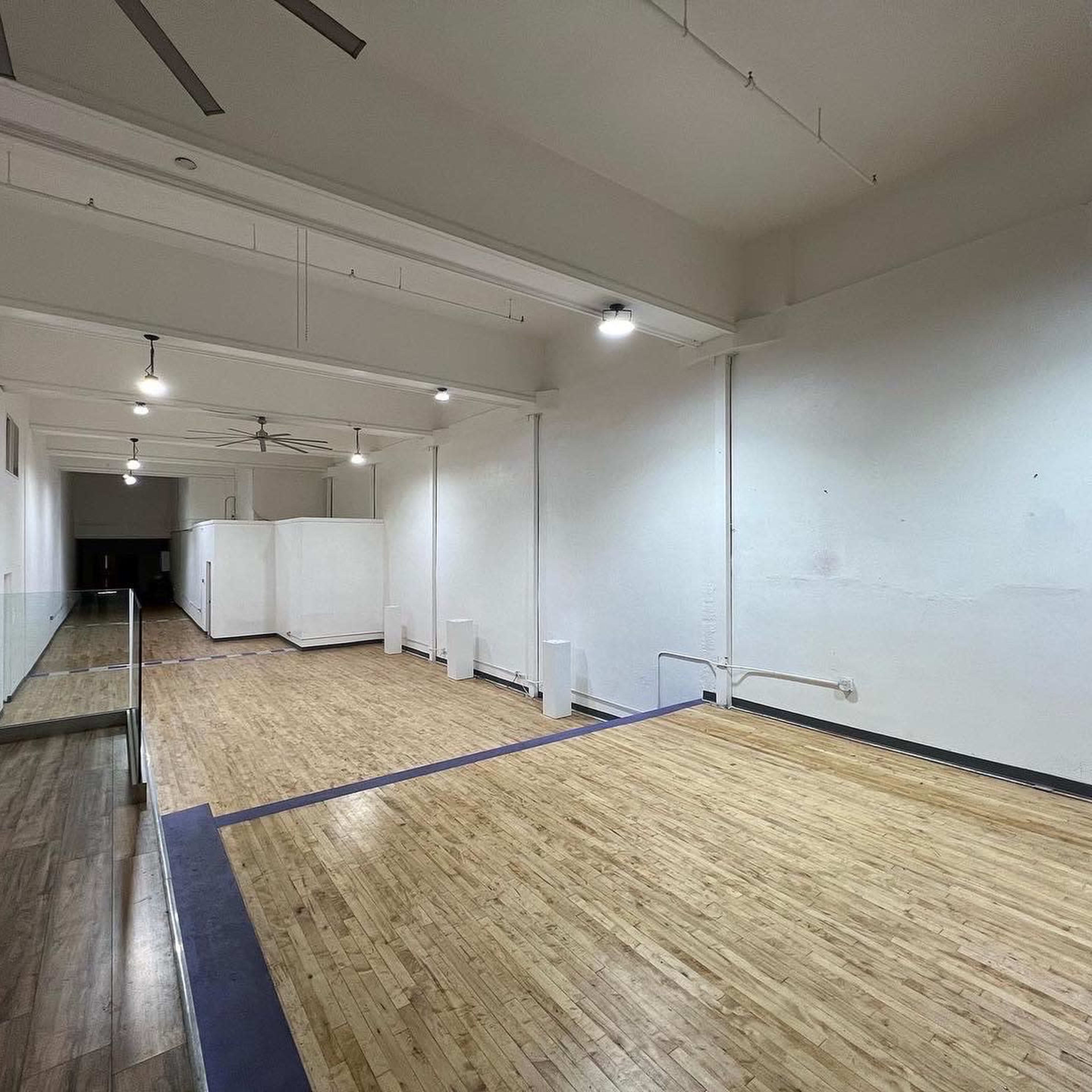 A spacious, empty room with wooden flooring and white walls, featuring ceiling fans and minimal furnishings.