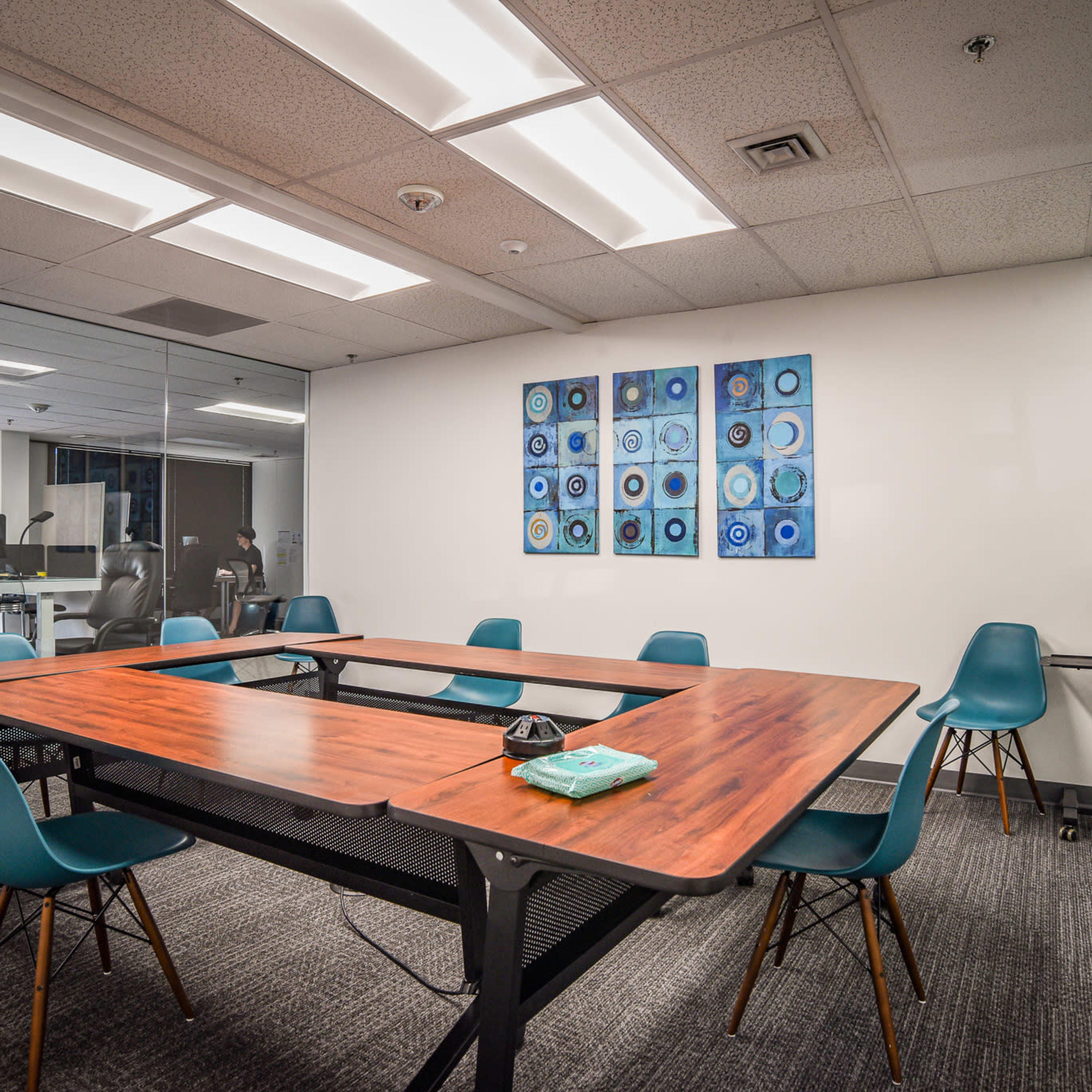 A modern conference room with a wooden table surrounded by teal chairs, and a large wall with framed blue artwork.
