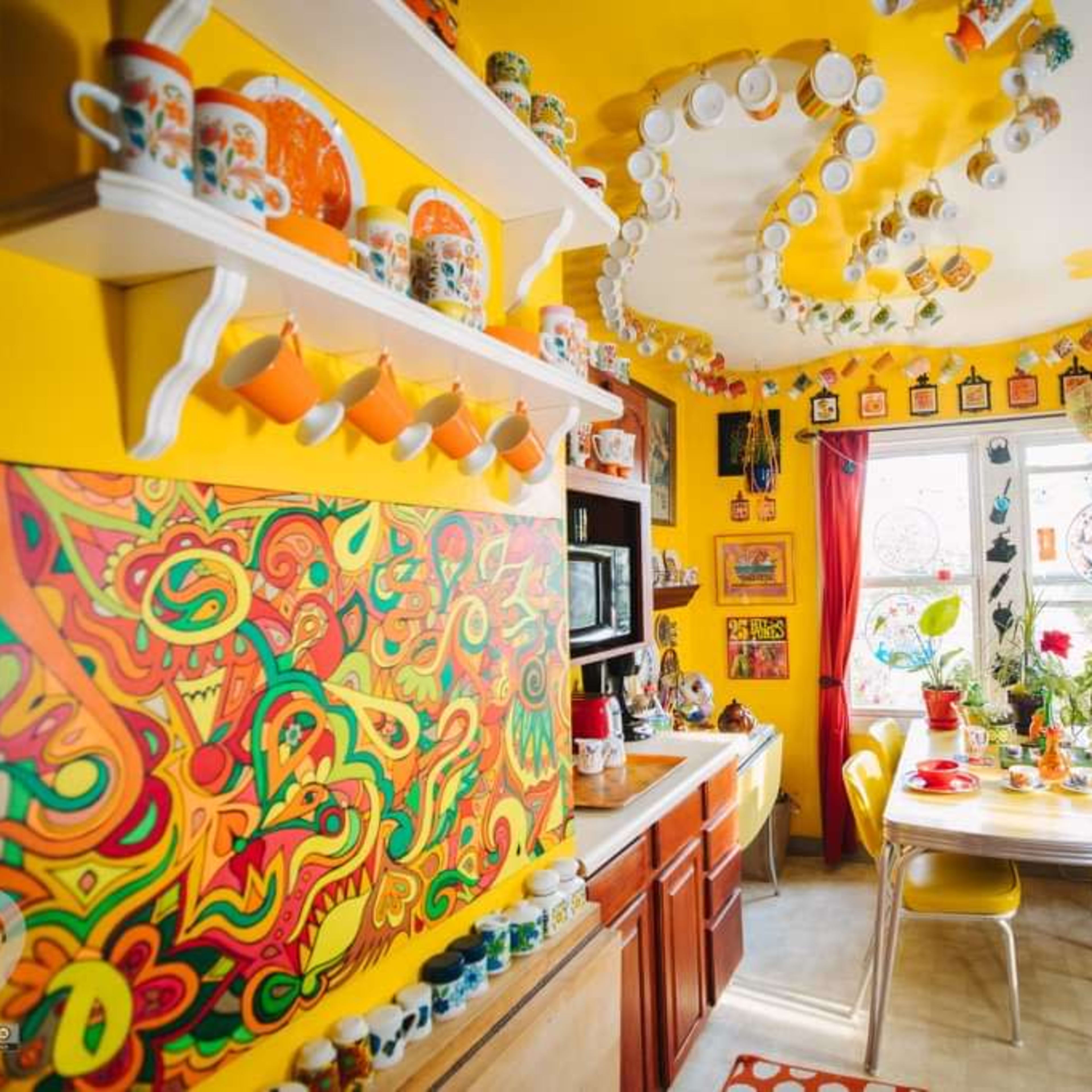 The kitchen features bright yellow walls adorned with colorful artwork and decorative cups, a table set for a meal, and a variety of kitchenware displayed on shelves.
