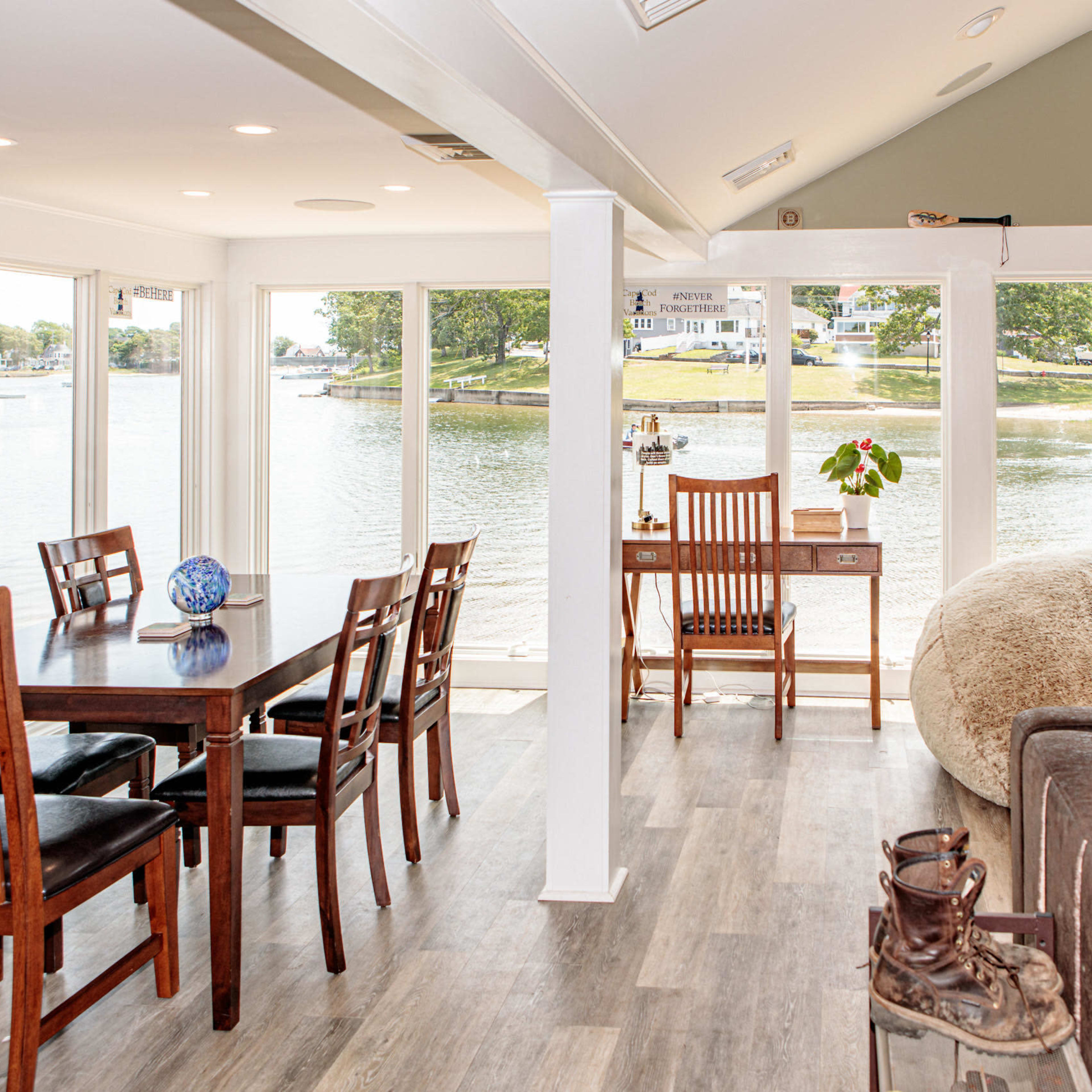 A bright, open living space with a dining table and chairs, a desk by the window, and a view of the water outside.