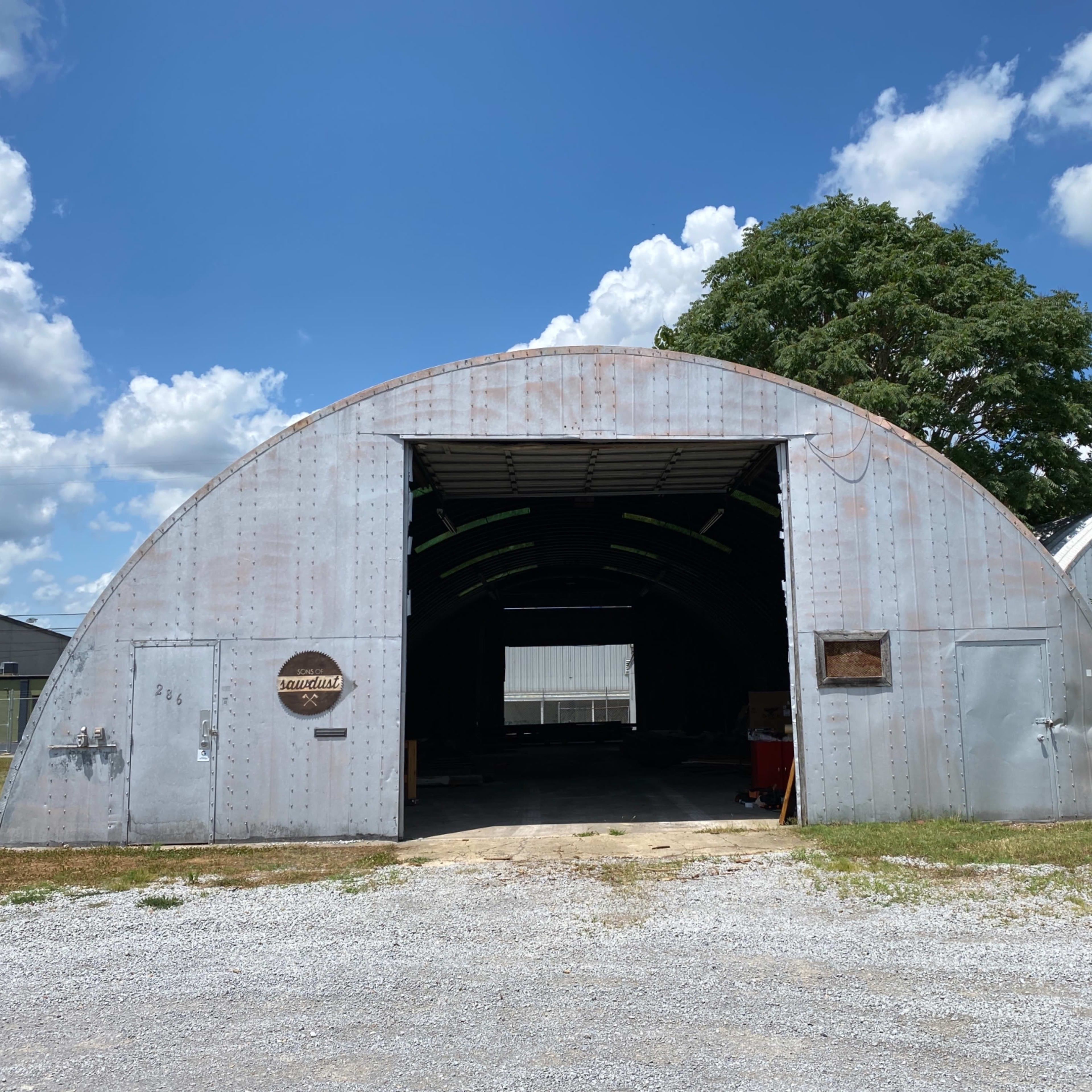 A large, corrugated metal structure with an arched roof and an open entrance, surrounded by gravel and green vegetation under a cloudy blue sky.
