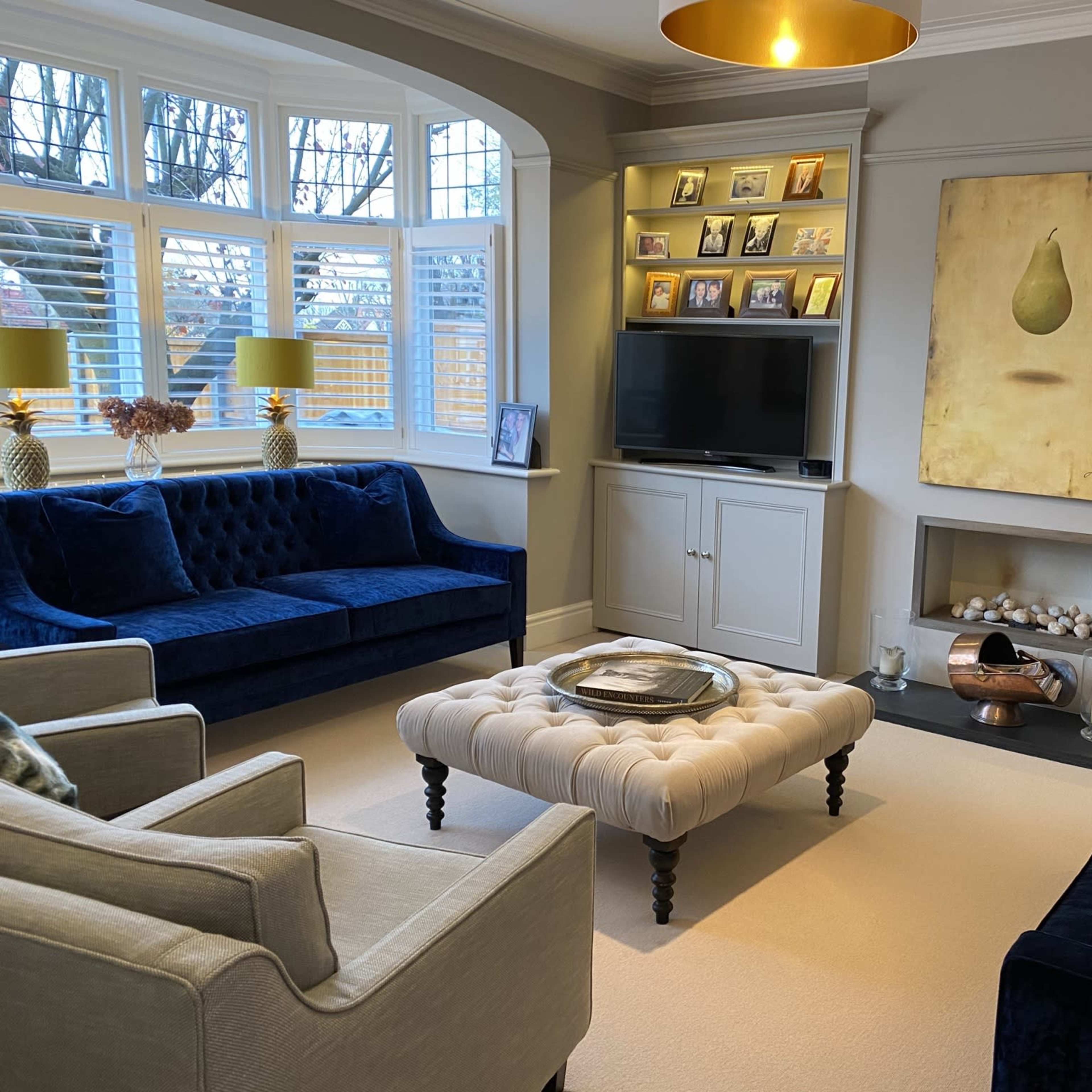 A modern living room features two blue velvet sofas, a tufted ottoman, a television, and a cream-colored area rug, with a wall art piece and shelves displaying photos and decorative items.
