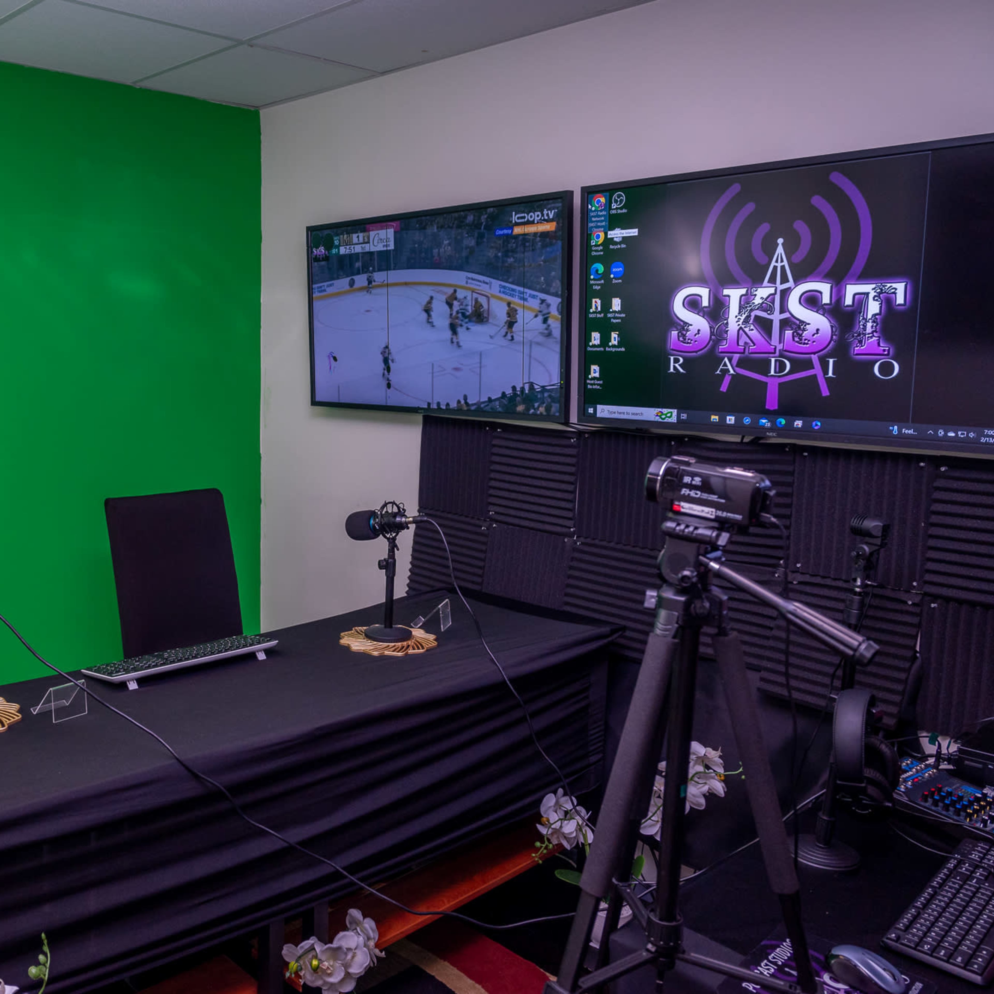 A recording studio with a green screen, two microphones, a desktop computer, and televisions displaying a sports game and a logo for a radio station.